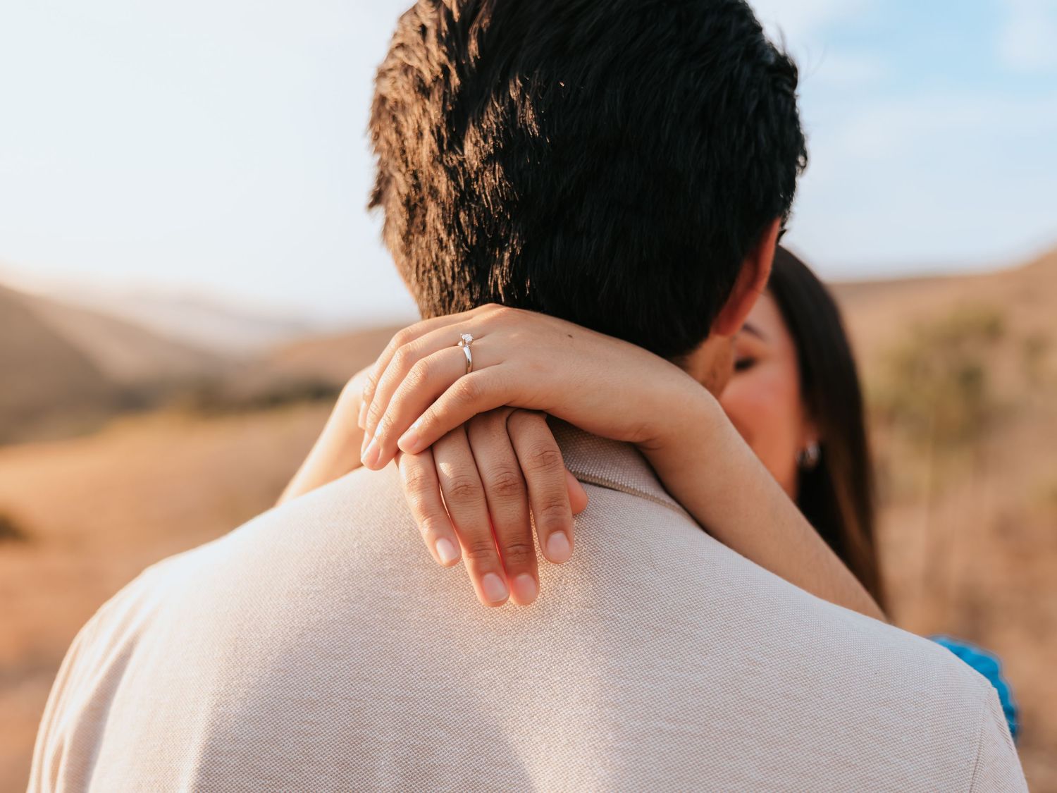 Couple embracing outdoors, woman with engagement ring resting hand on man's shoulder against scenic landscape.