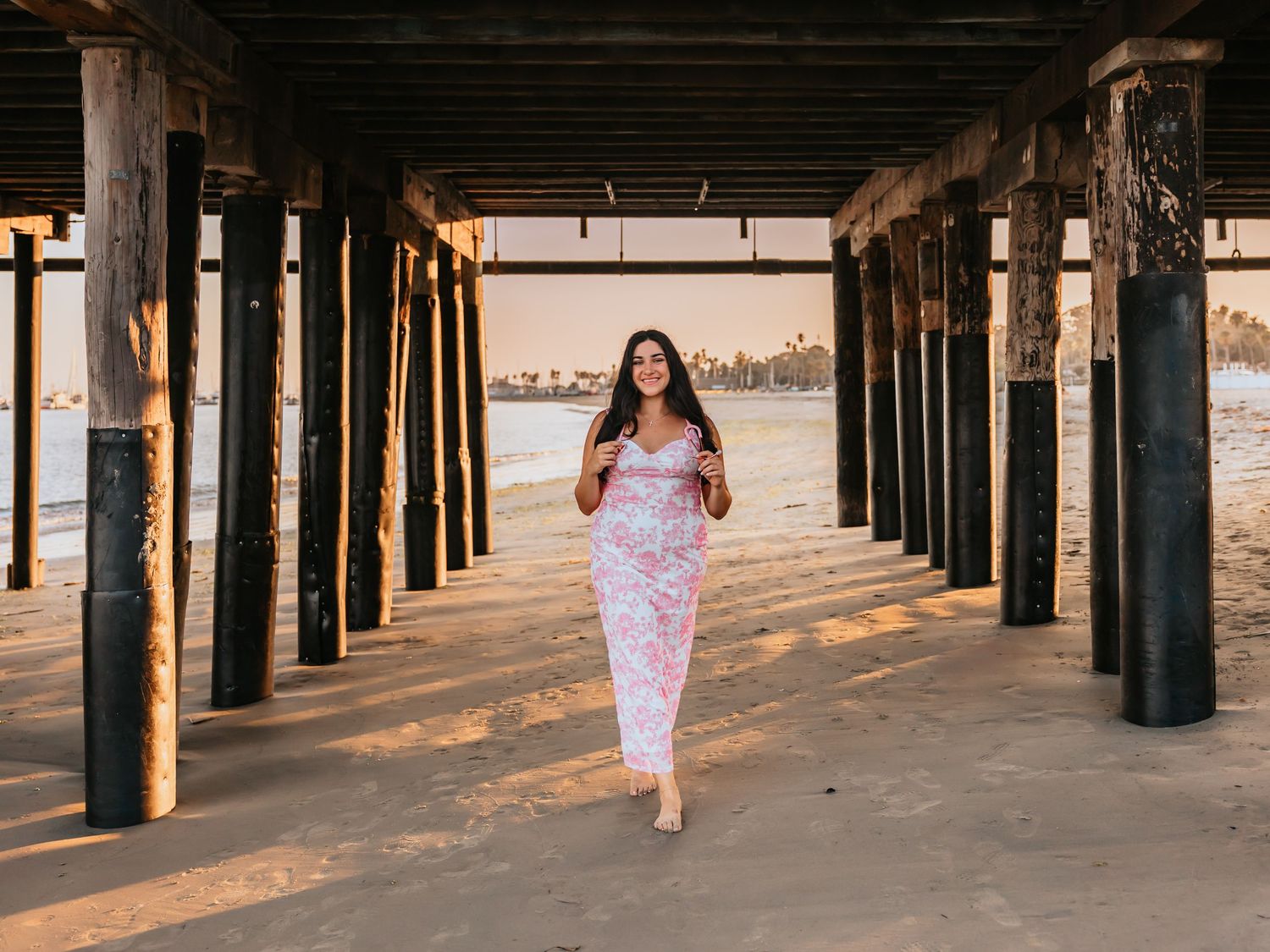 Woman in floral maxi dress walking under a beach pier at golden hour on sandy shore.
