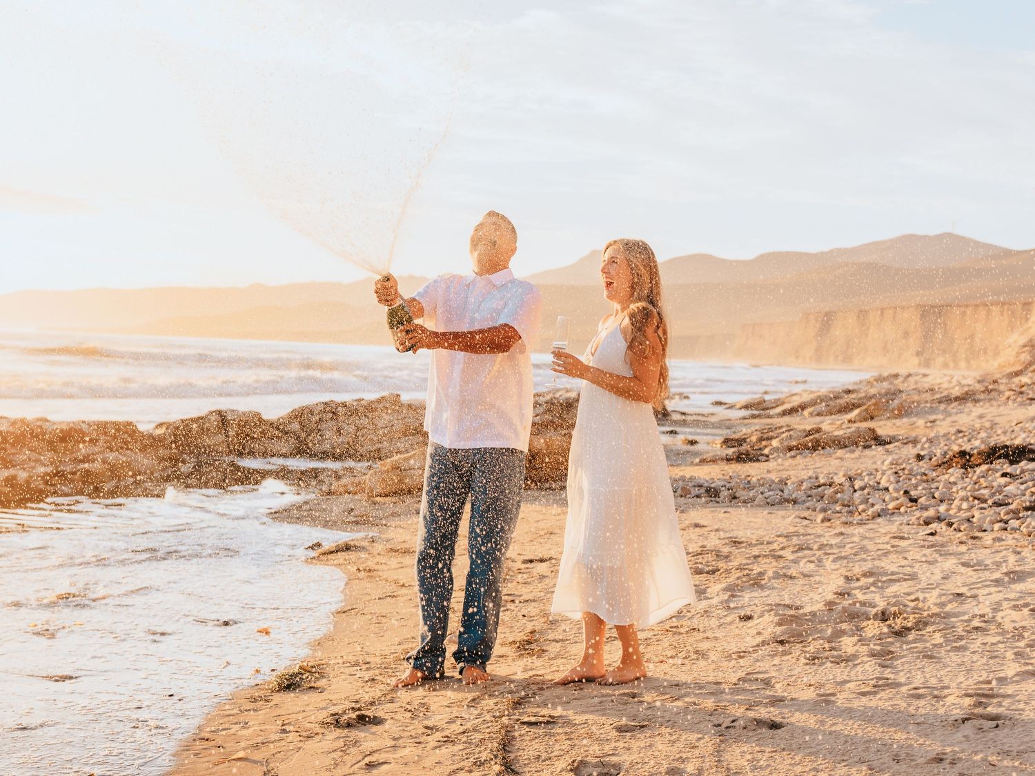 Couple celebrating on a sunny beach, popping champagne with ocean waves and golden cliffs in the background.