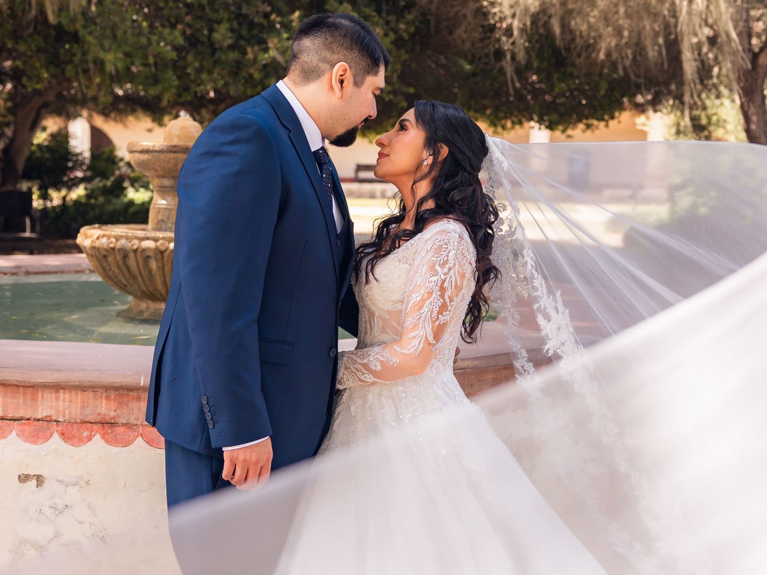 A bride and groom share a romantic kiss outdoors near a fountain, her veil flowing gracefully in the breeze.