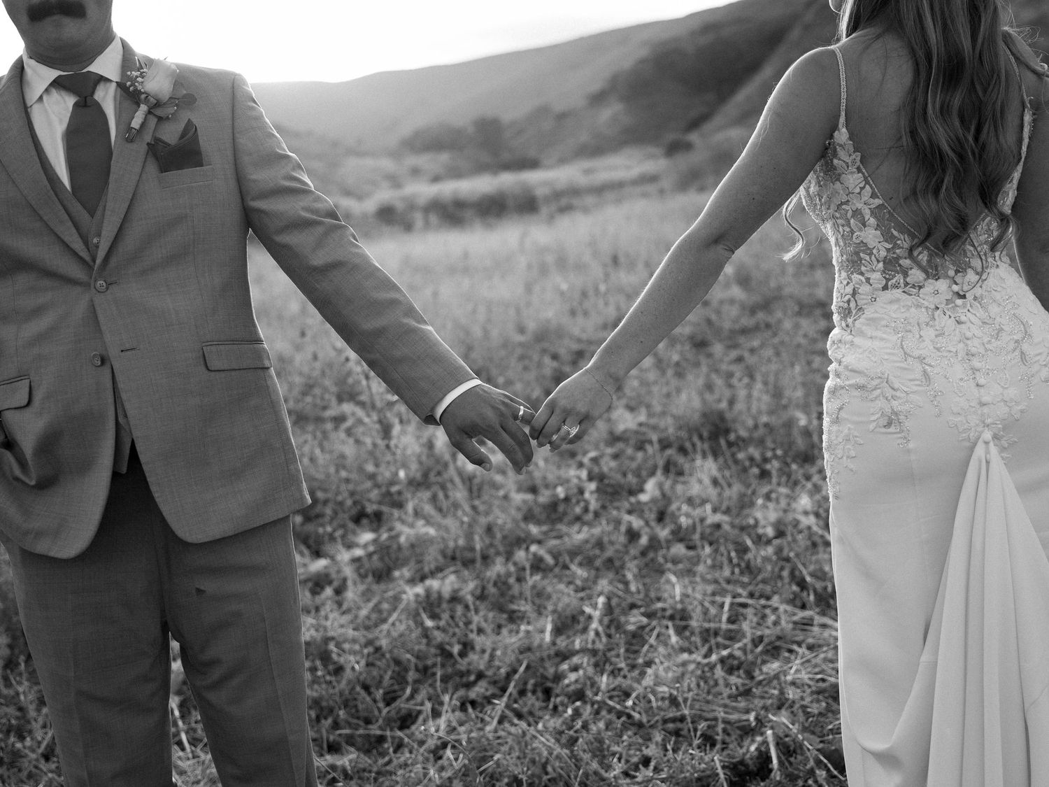 Black and white photo of a bride and groom holding hands in a field, wearing wedding attire.