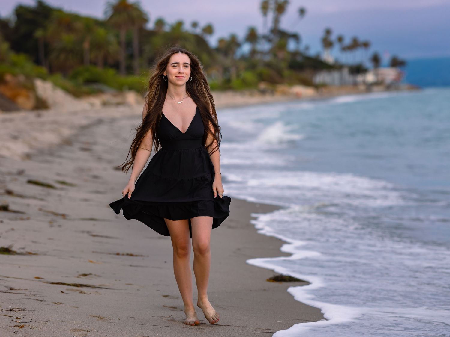 Woman in black dress walking barefoot along tropical beach with palm trees and ocean waves at dusk.