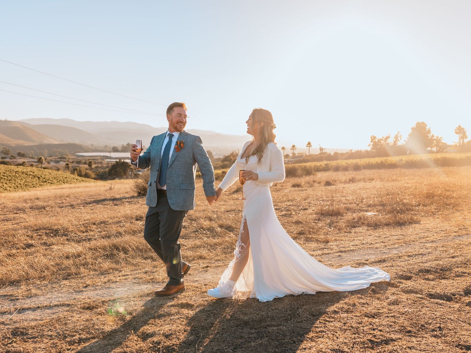 A couple in wedding attire walking hand in hand across golden hills at sunset with a flowing white dress trailing behind.
