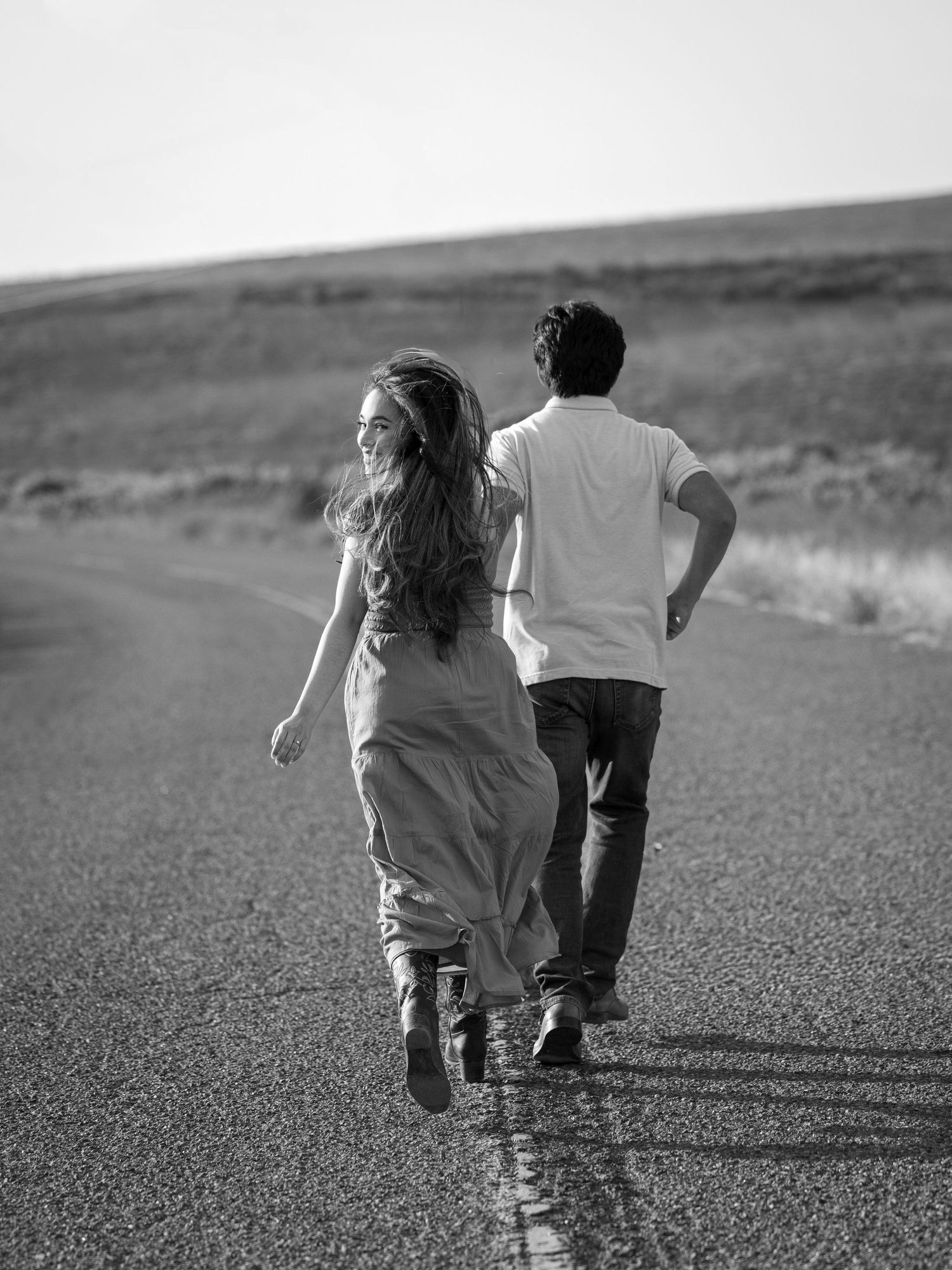 Black and white photo of a couple walking down an open road, woman smiling with long hair, man in white t-shirt.