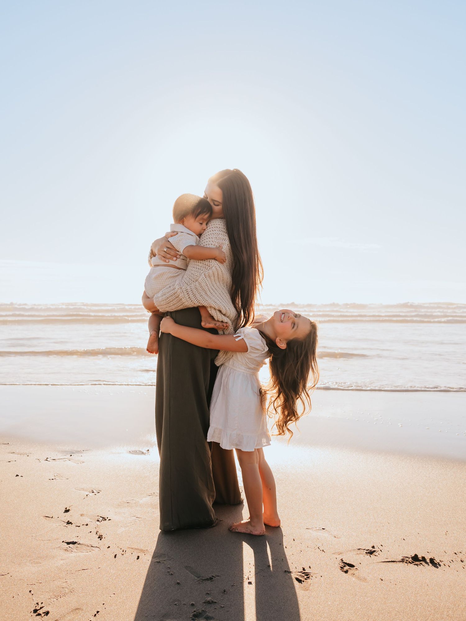 A mother holds her baby while her young daughter hugs her on a sunny beach with ocean waves in the background.