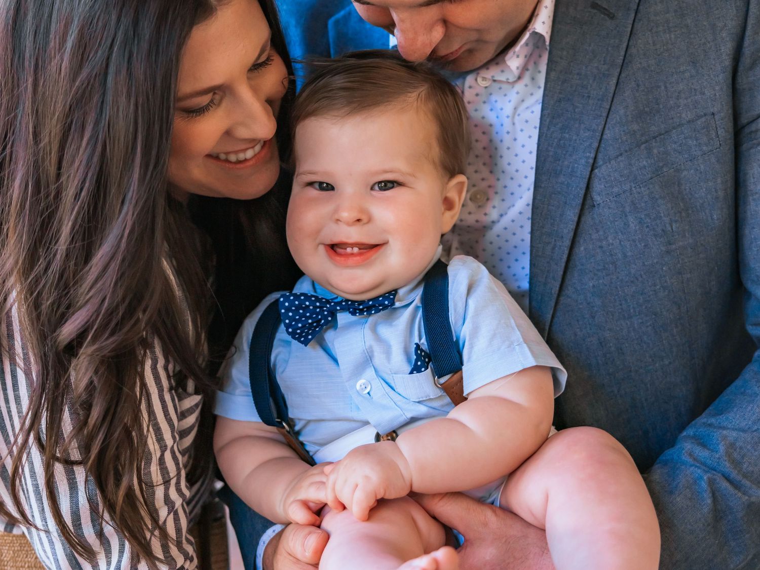 A smiling baby wearing a blue bowtie sits between two adults in a casual portrait setting.
