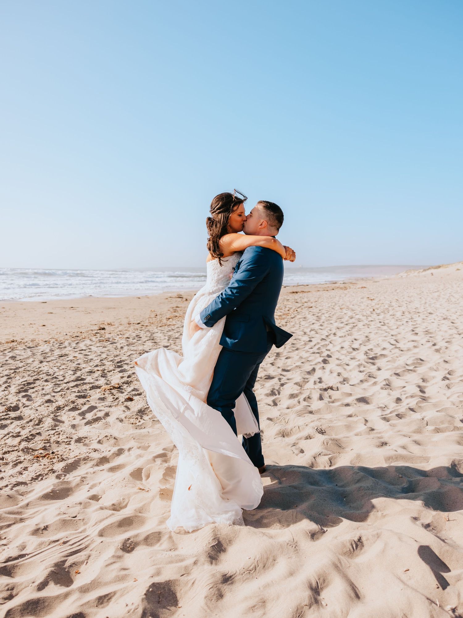 Groom lifts bride in white wedding dress on sandy beach, sharing a romantic kiss with ocean waves in the background.