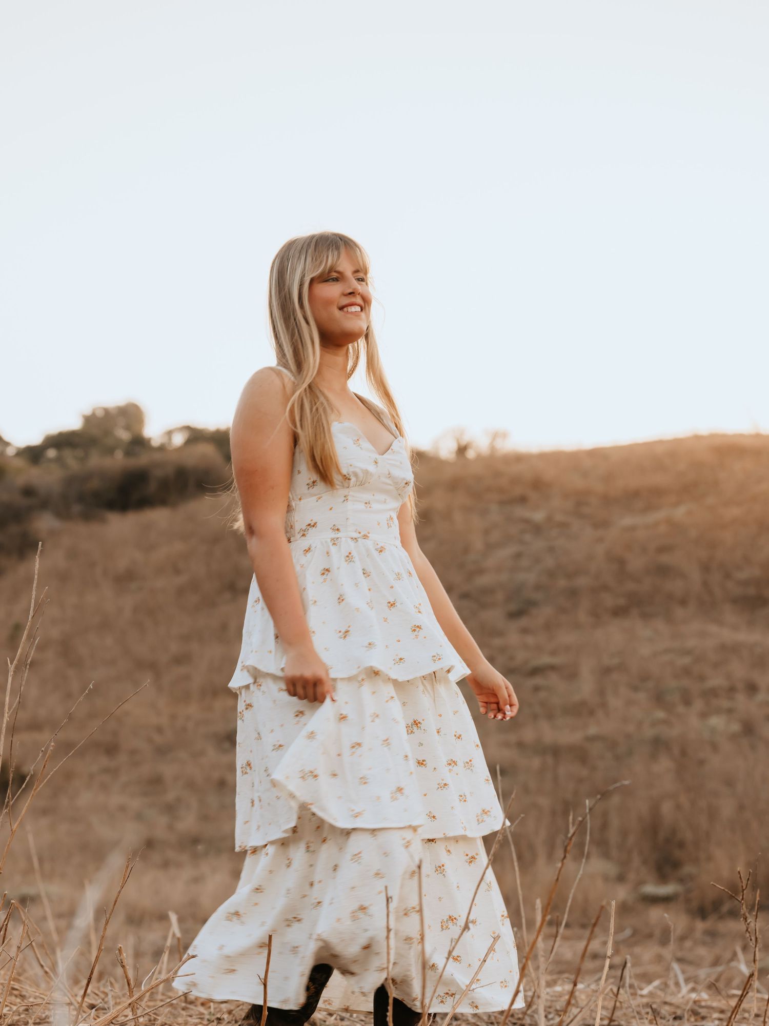Blonde woman in a white floral tiered maxi dress standing in a golden field during sunset.
