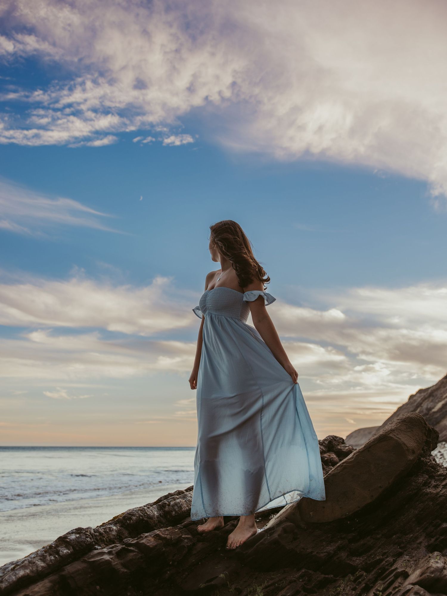 Woman in flowing light blue off-shoulder gown standing on rocky coastline at sunset with dramatic cloudy sky.