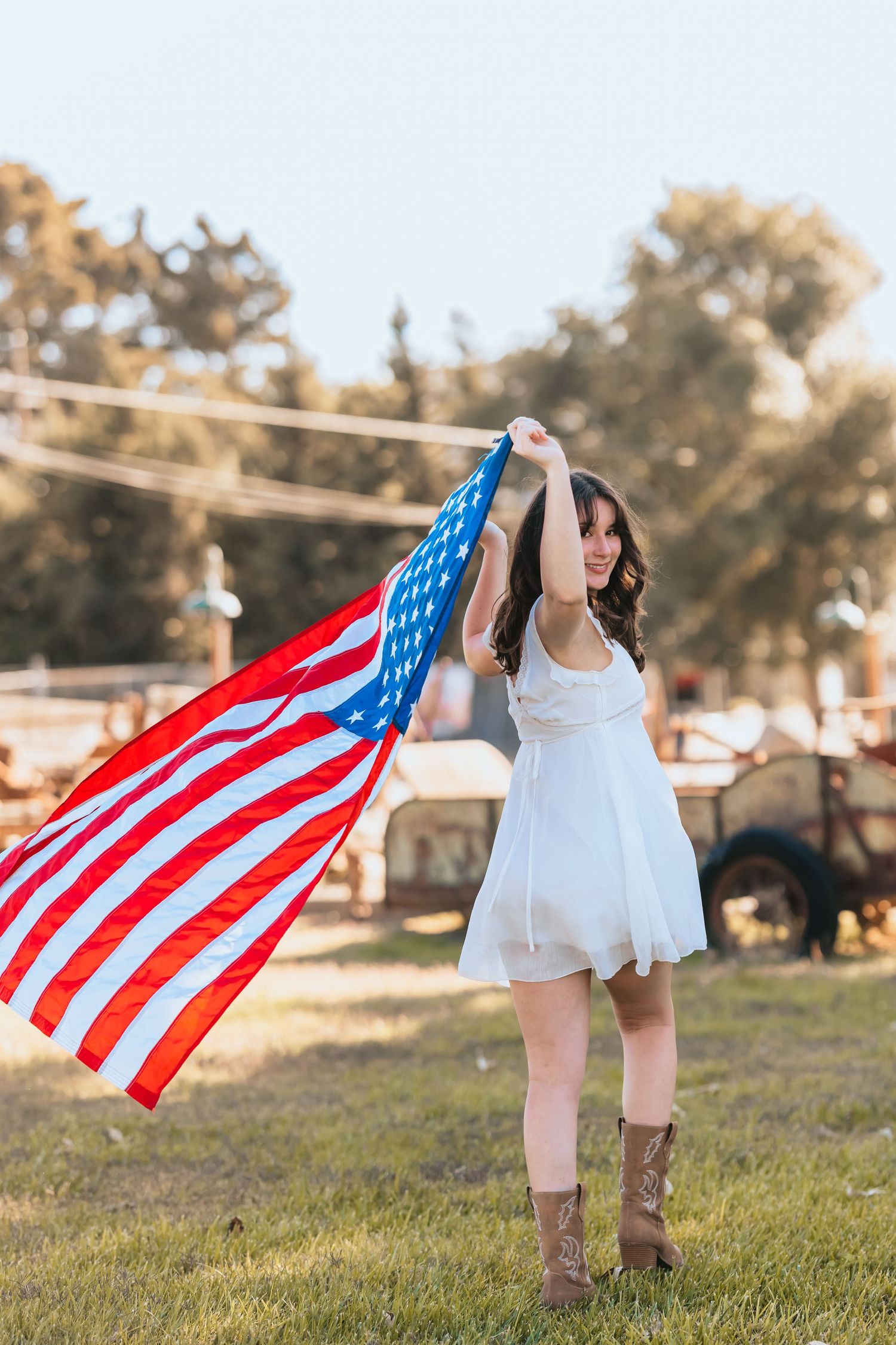 Woman in white dress raising American flag in sunny field wearing brown cowboy boots.
