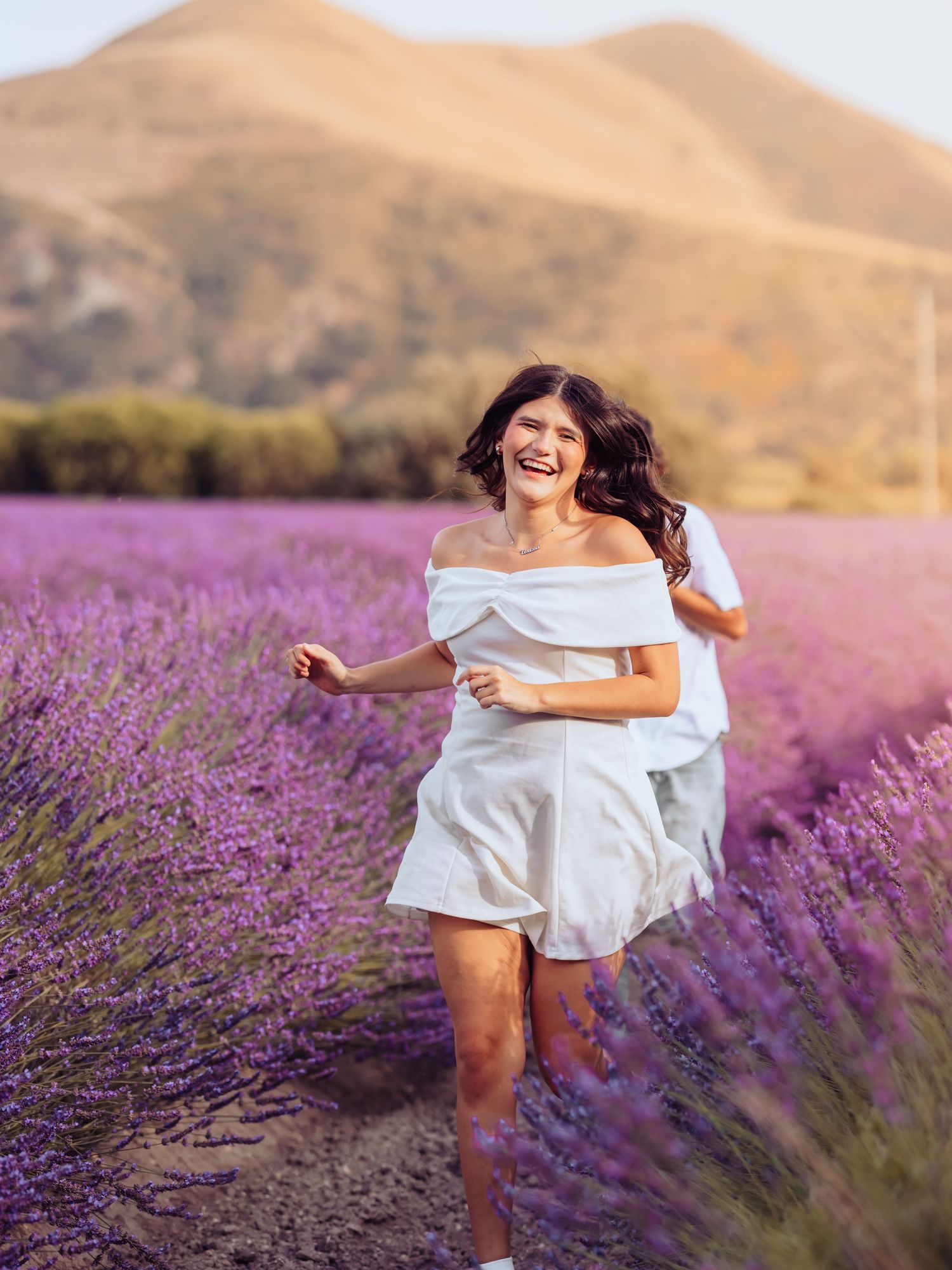 A joyful woman in a white off-shoulder dress runs through a vibrant purple lavender field with golden hills behind her.