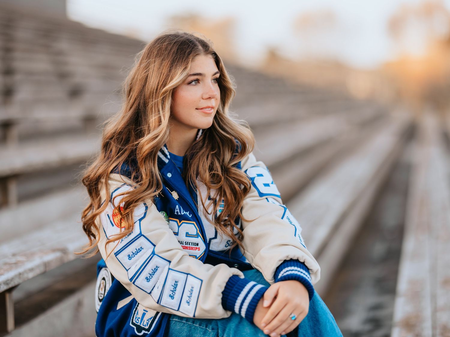 A person in a blue and white varsity jacket sitting on bleachers during golden hour.