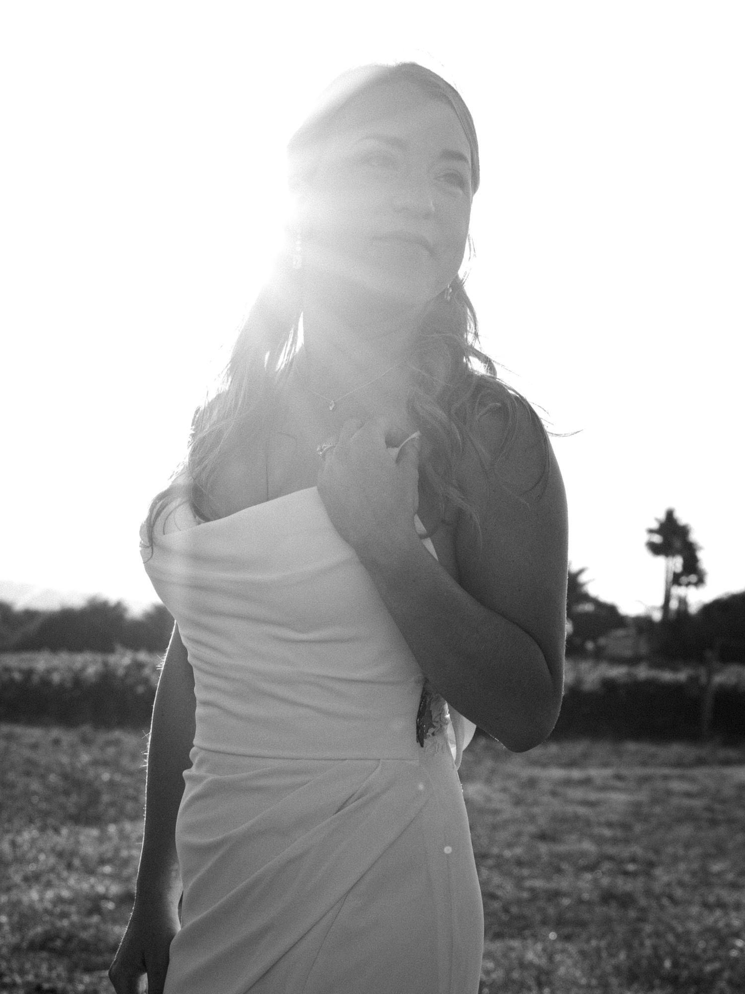 Black and white photo of a woman in a white strapless dress with sun flare behind her in an outdoor field.
