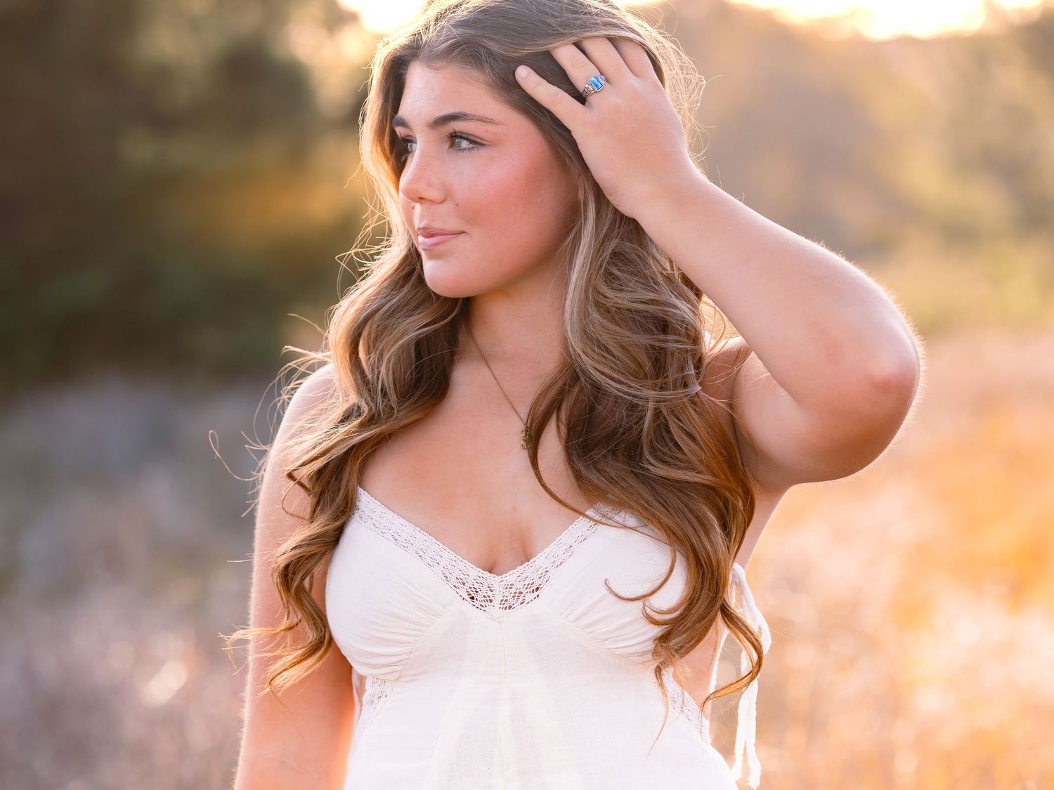 A woman with long wavy brown hair poses outdoors in a white dress during golden hour sunset.
