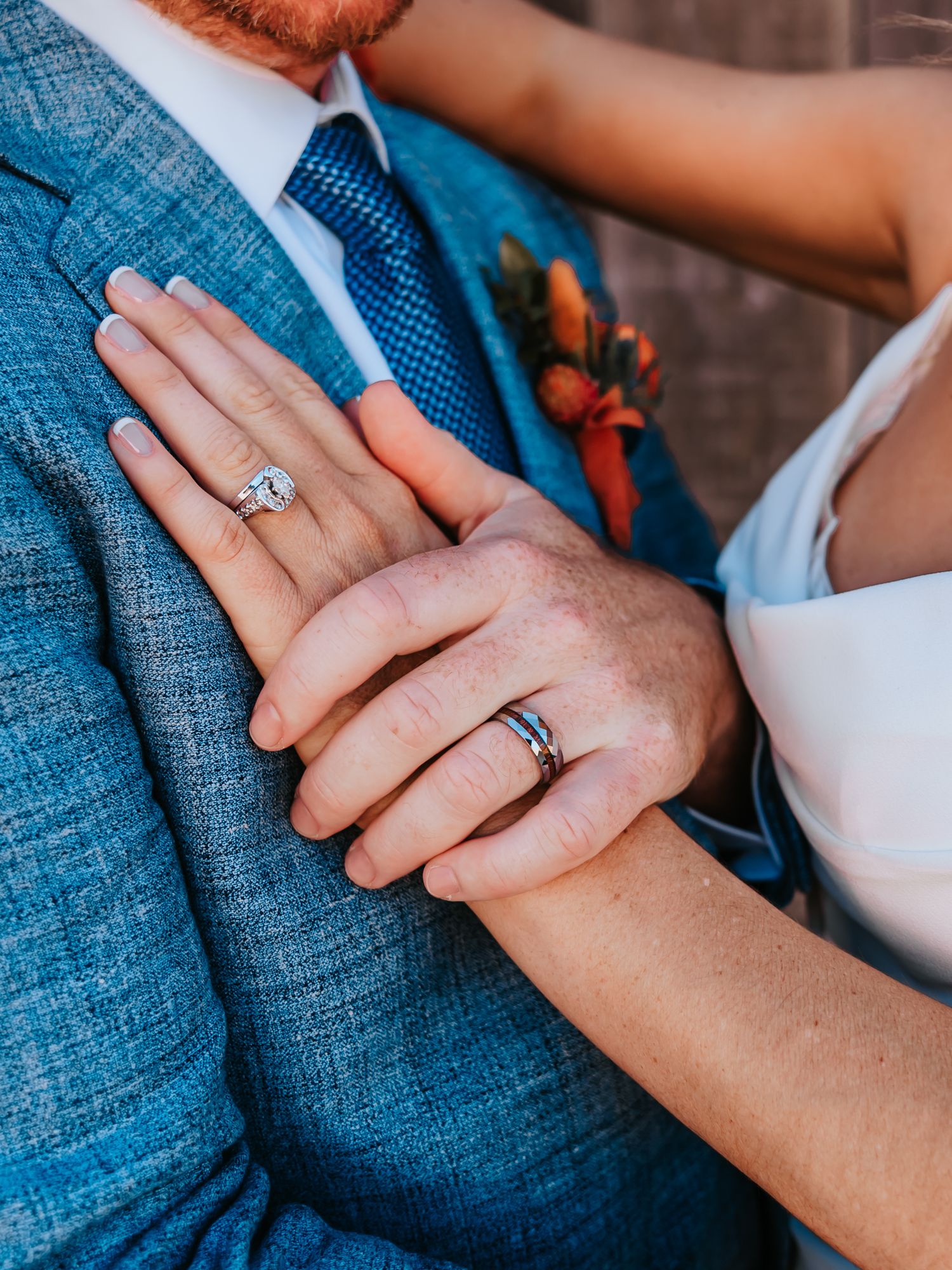 Close-up of bride and groom's hands showing wedding rings, groom in blue suit with floral boutonniere.