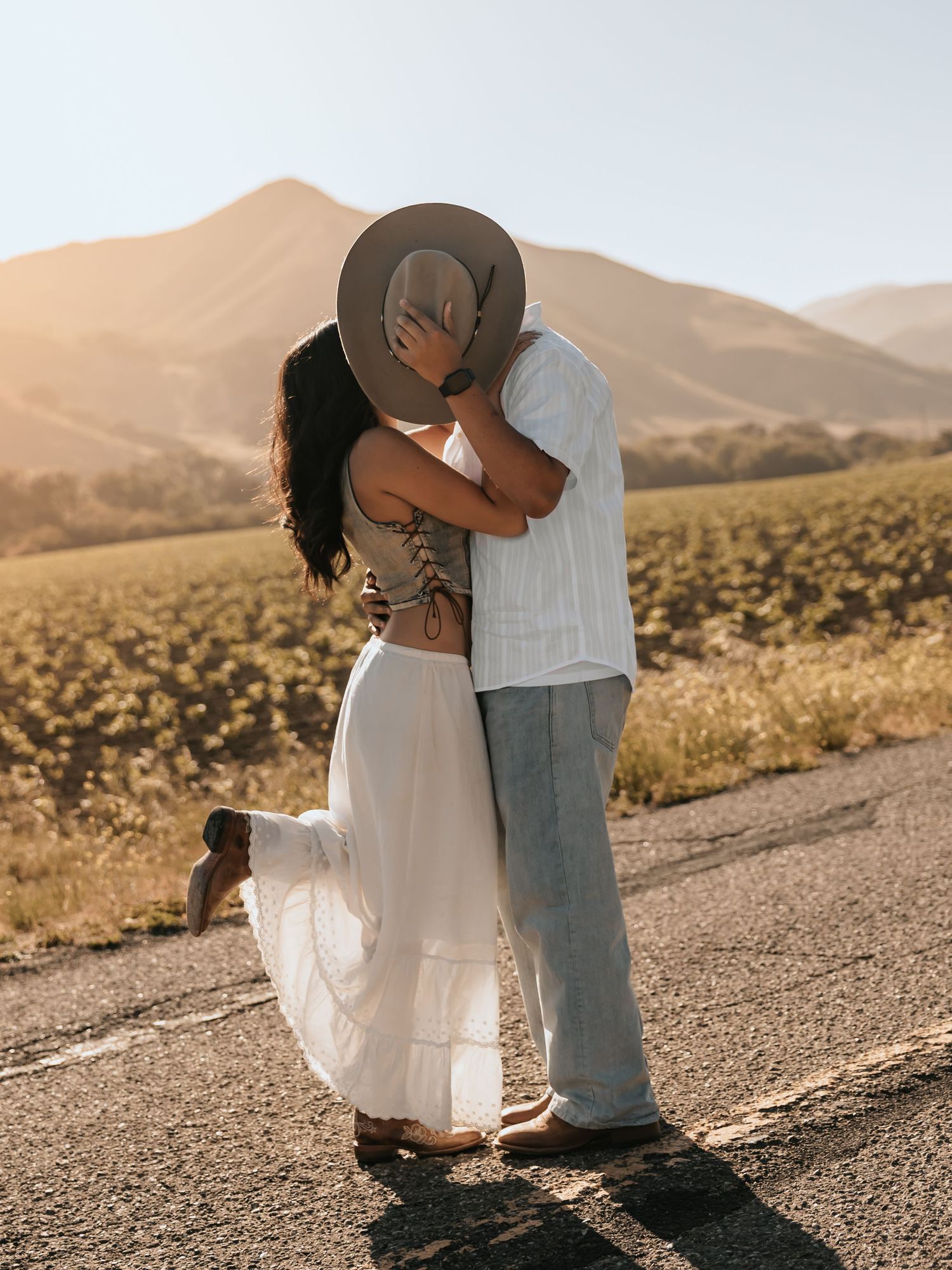Couple sharing a romantic kiss on a rural road with golden hills and green fields in the background at sunset.