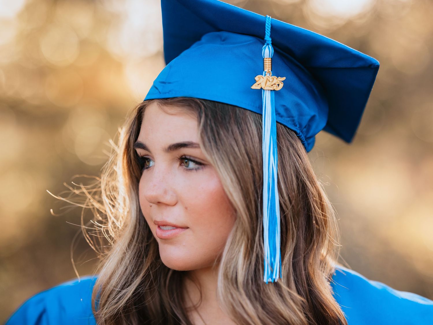 Graduate in bright blue cap and gown with gold tassel looks off to the side during outdoor graduation photo session.
