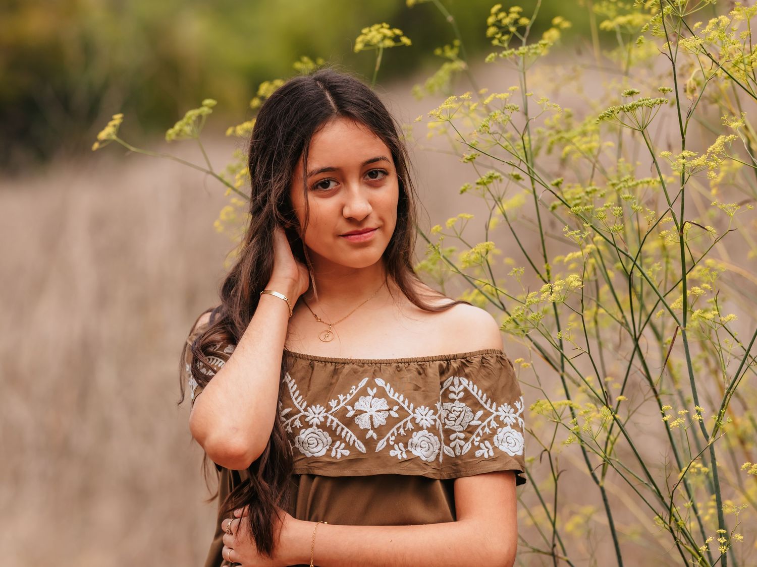 Young woman in embroidered off-shoulder dress poses gracefully among wild green plants in a natural outdoor setting.