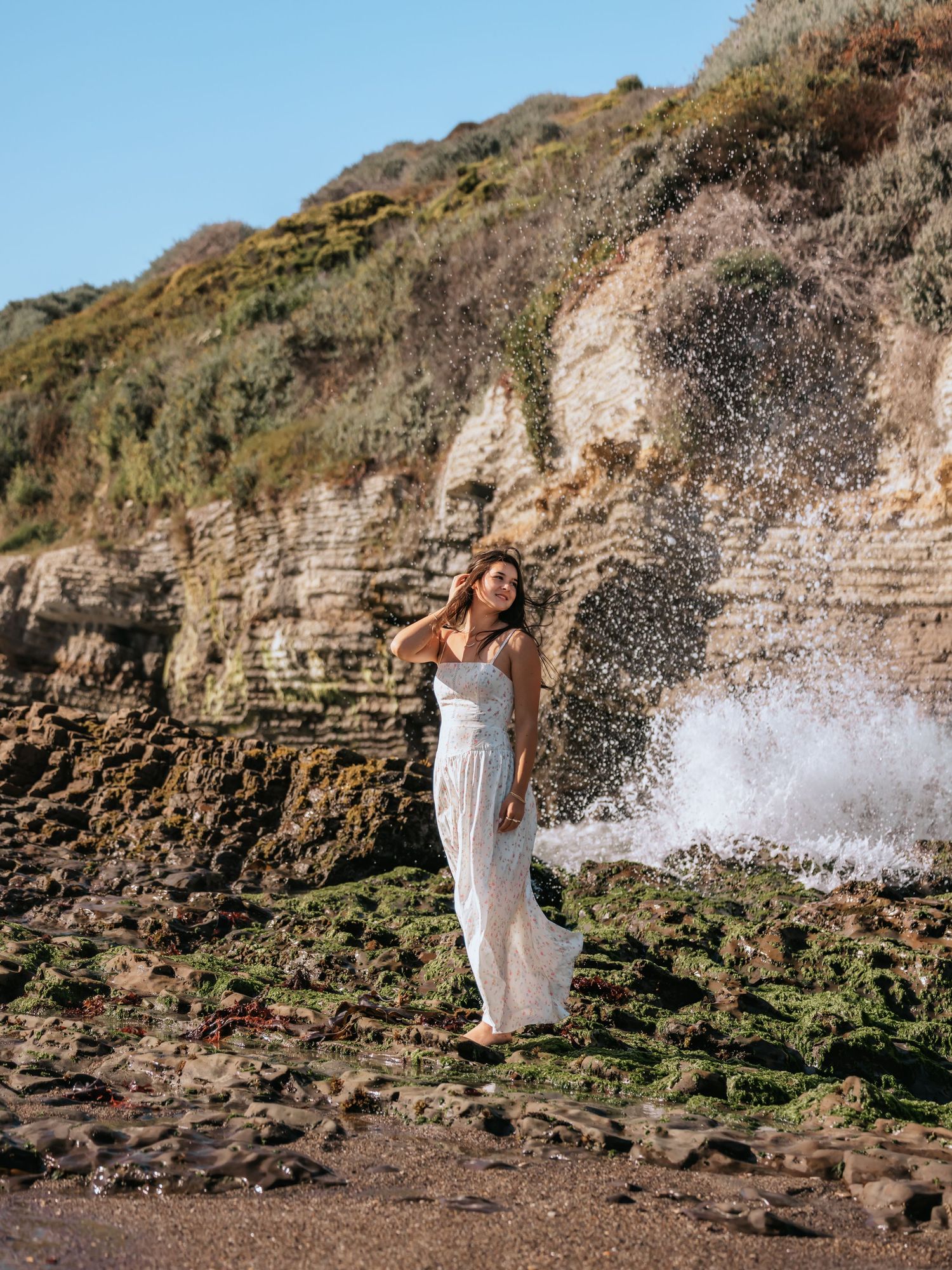 Woman in white dress standing on rocky shore with crashing waves and cliffside backdrop.