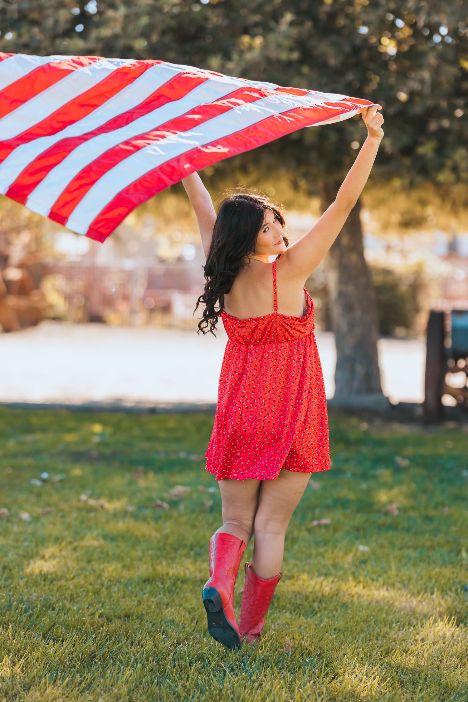 Woman in red dress raising American flag overhead in sunny park wearing pink cowboy boots.
