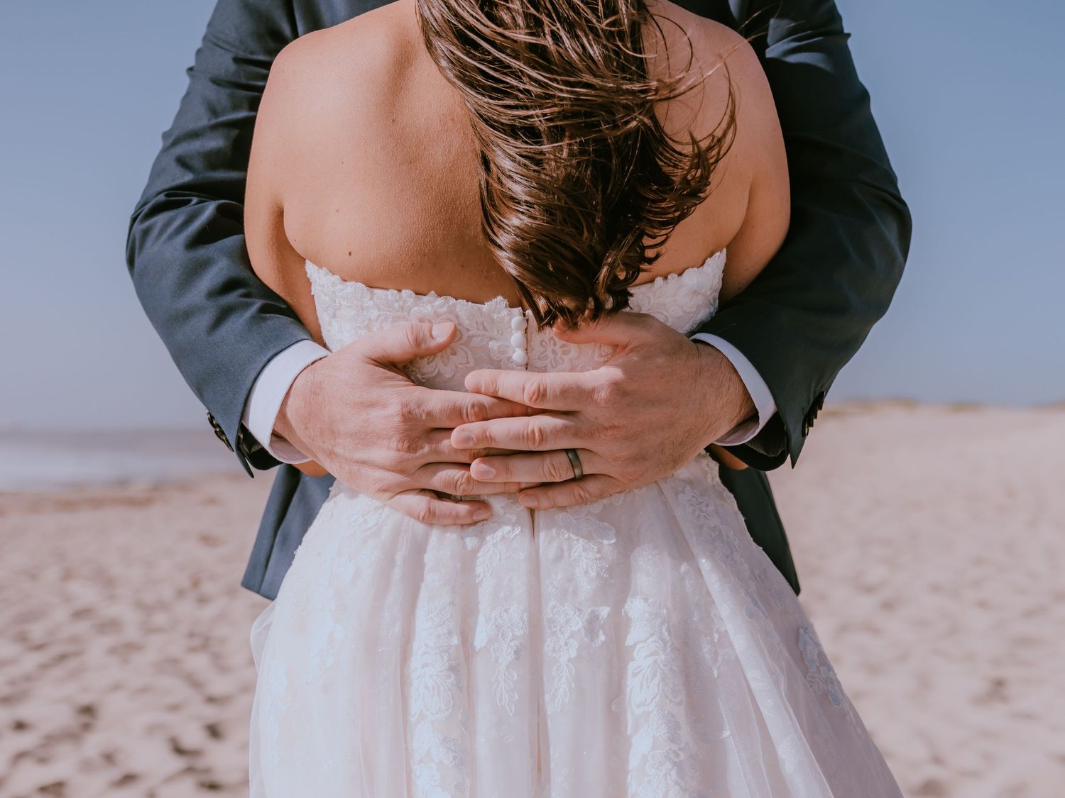 A couple embraces on a sandy beach with the bride wearing a white lace dress and groom in a dark suit.