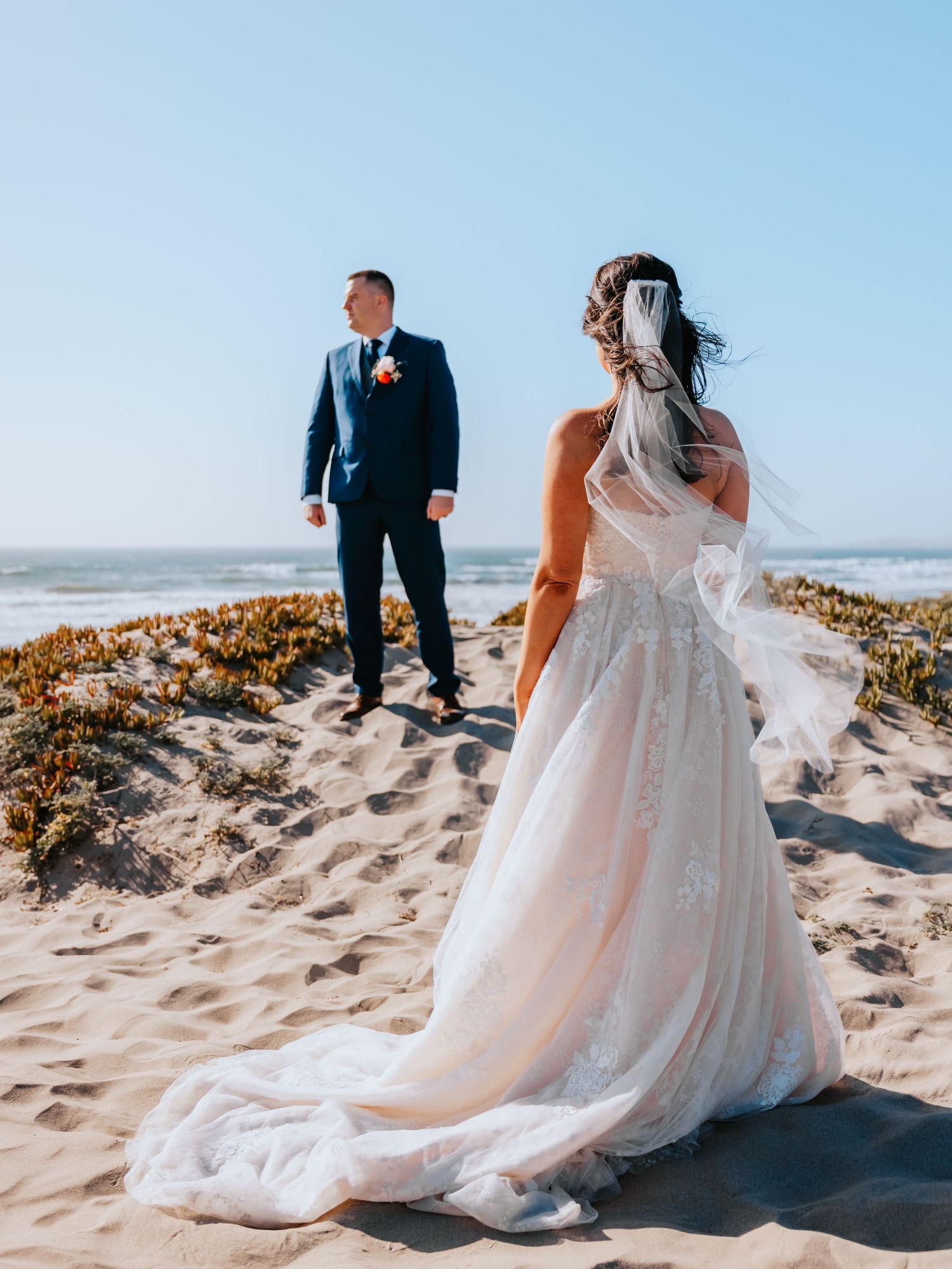 Bride in lace gown with veil and groom in navy suit share a romantic beach moment on sandy shores.