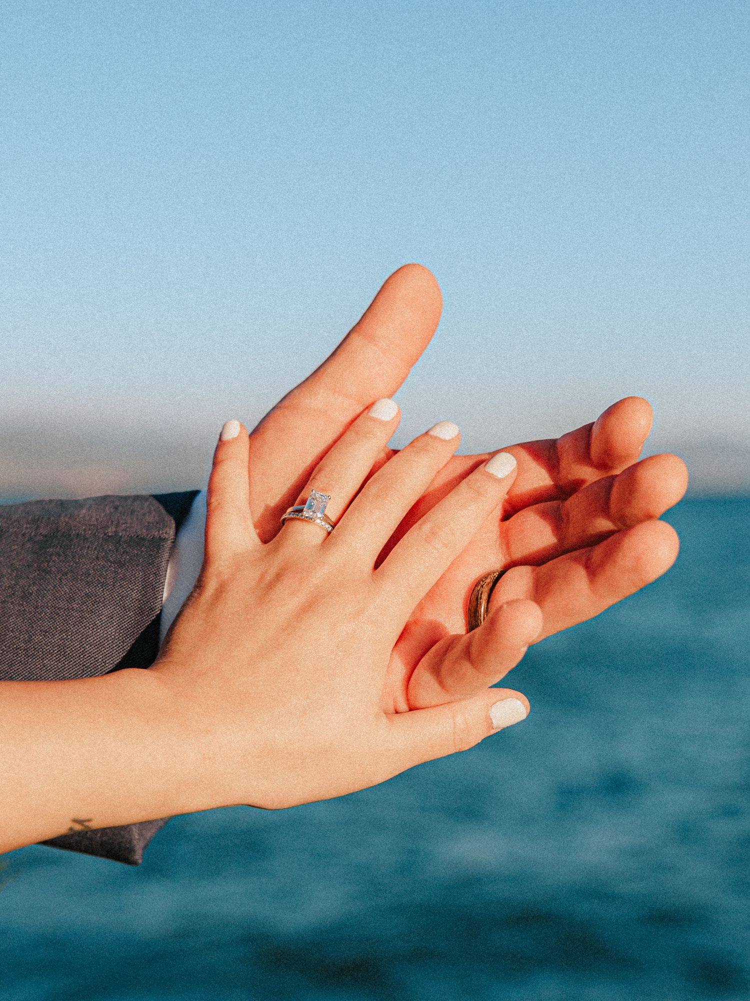 Two hands raised against blue sky and ocean, one wearing a diamond engagement ring and white nail polish.