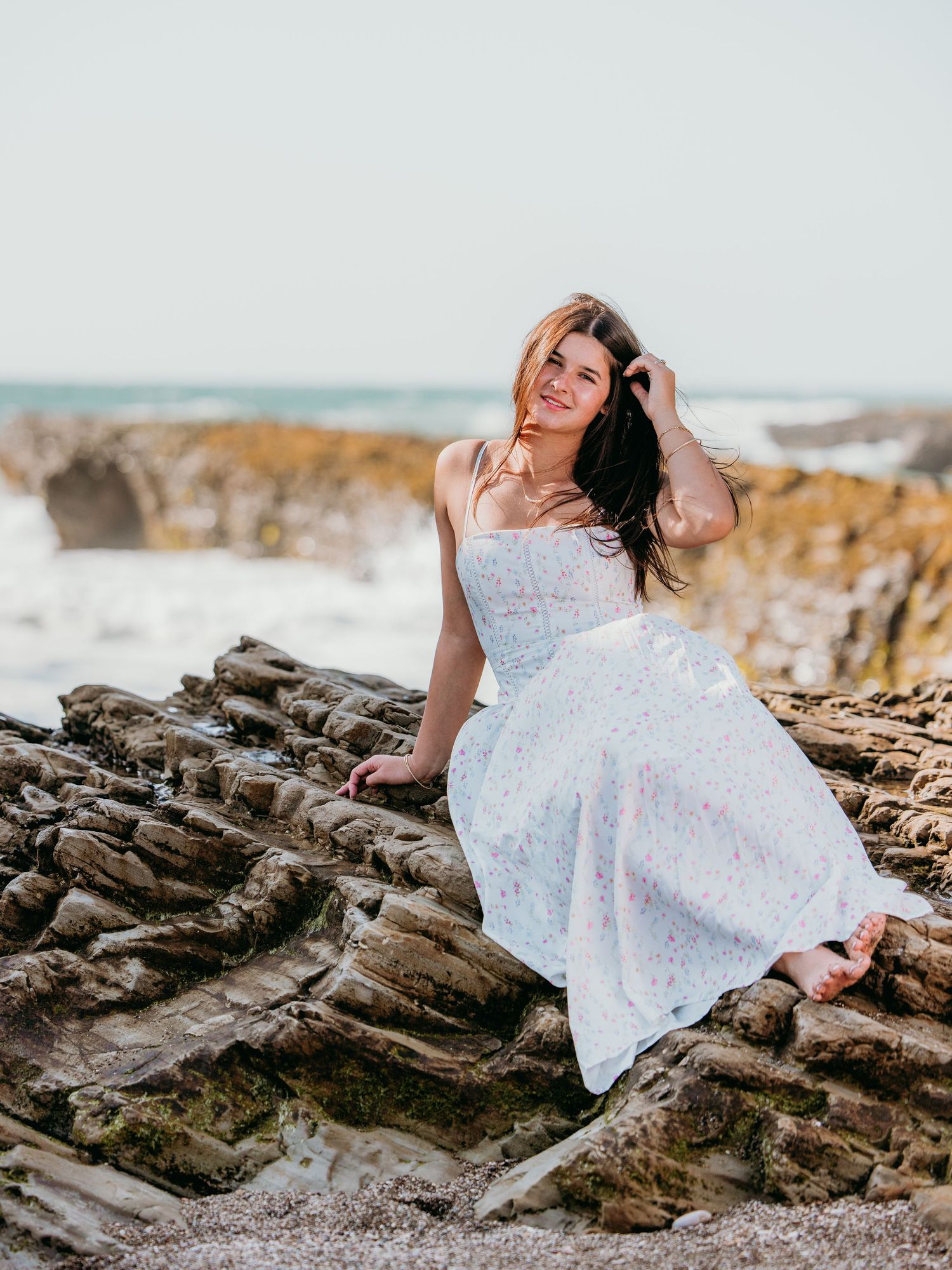 A young woman in a white floral maxi dress sits on rocky coastal cliffs with ocean waves in the background.