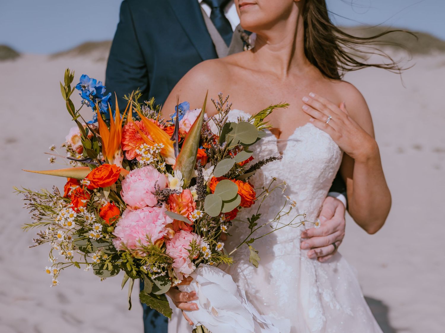 A vibrant wedding bouquet with orange, pink and blue flowers held against a white wedding gown on a beach.