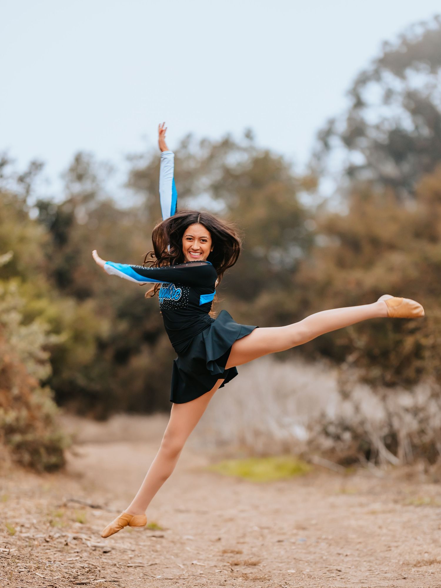 Dancer in black dress performs graceful leap against natural outdoor backdrop.
