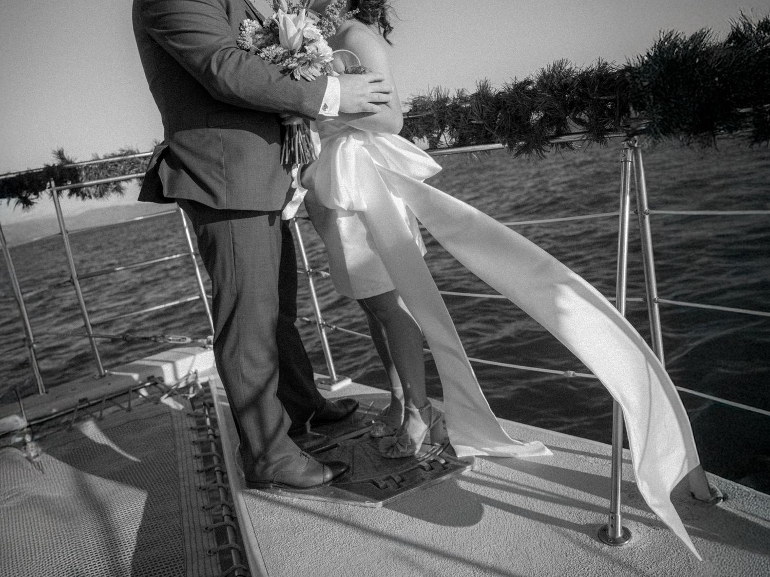 Black and white photo of a bride and groom embracing on a sailboat deck, with water and trees in the background.