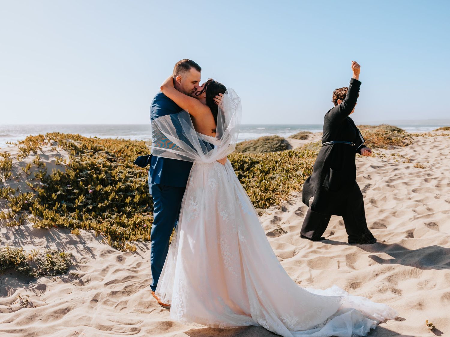 Bride and groom share a first kiss on a sandy beach while officiant celebrates behind them on a sunny day.