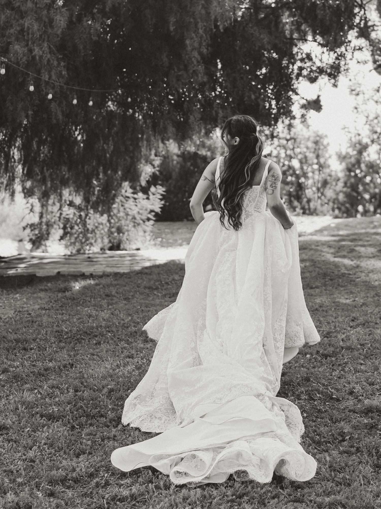 Black and white photo of a bride in a flowing lace wedding gown with a long train, standing on grass under willow trees.