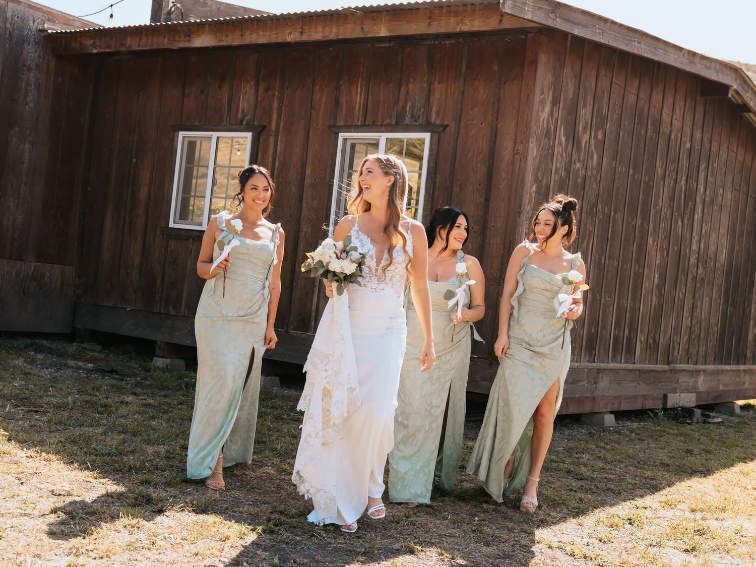Bride in white gown walks with four bridesmaids in sage green dresses outside a rustic wooden barn.