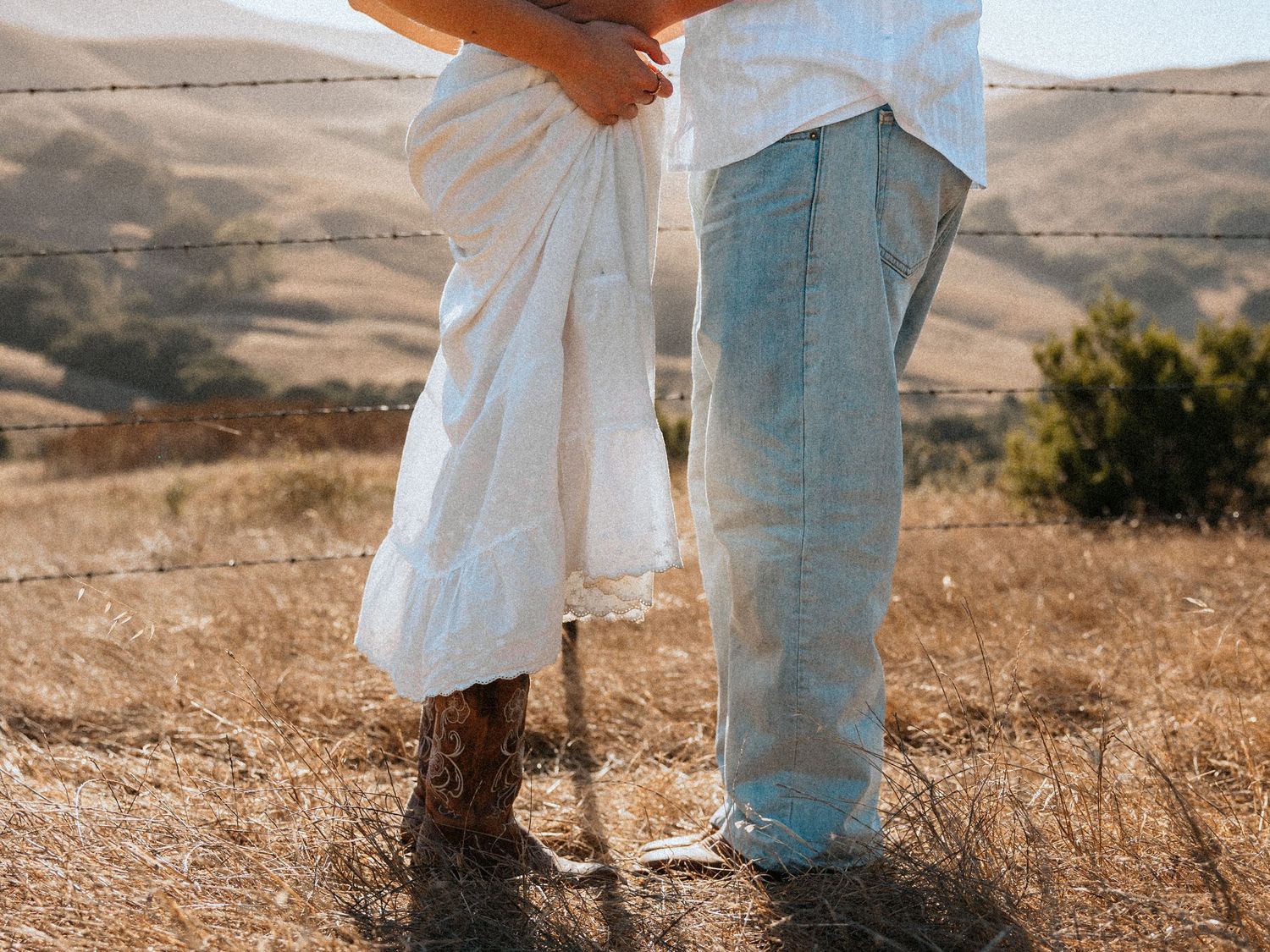 Couple standing close together in a sunny field, wearing white dress and light blue jeans with cowboy boots.