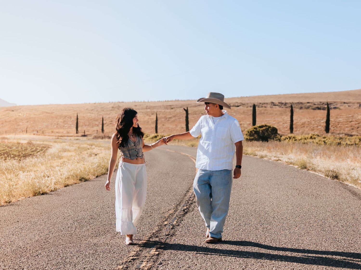 Couple holding hands while walking on a rural road surrounded by golden hills on a sunny day.