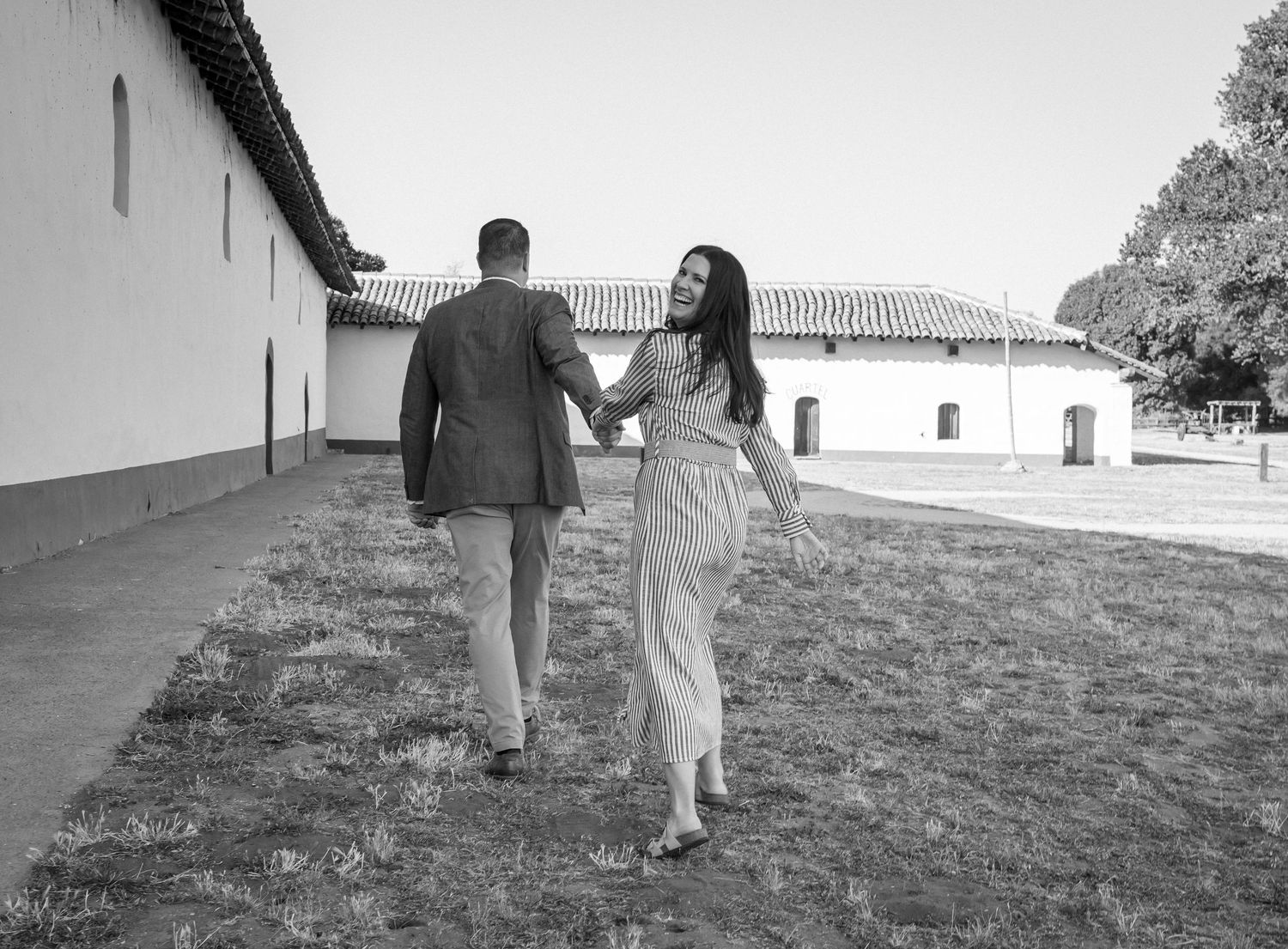 Black and white image of couple walking together holding hands on grassy path near white Spanish colonial style buildings.