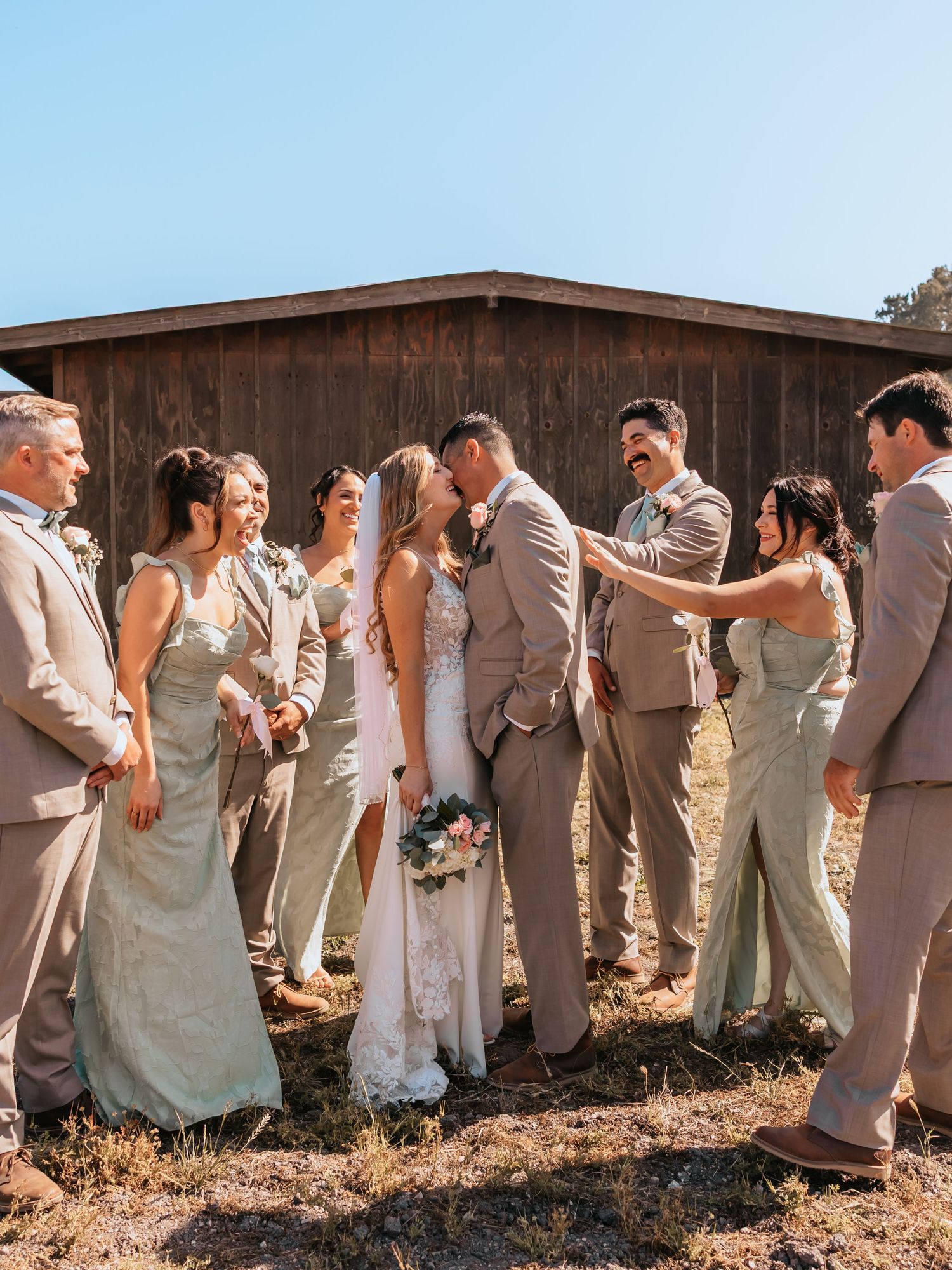 Wedding party kissing and celebrating outside rustic barn on sunny day in neutral and sage attire.