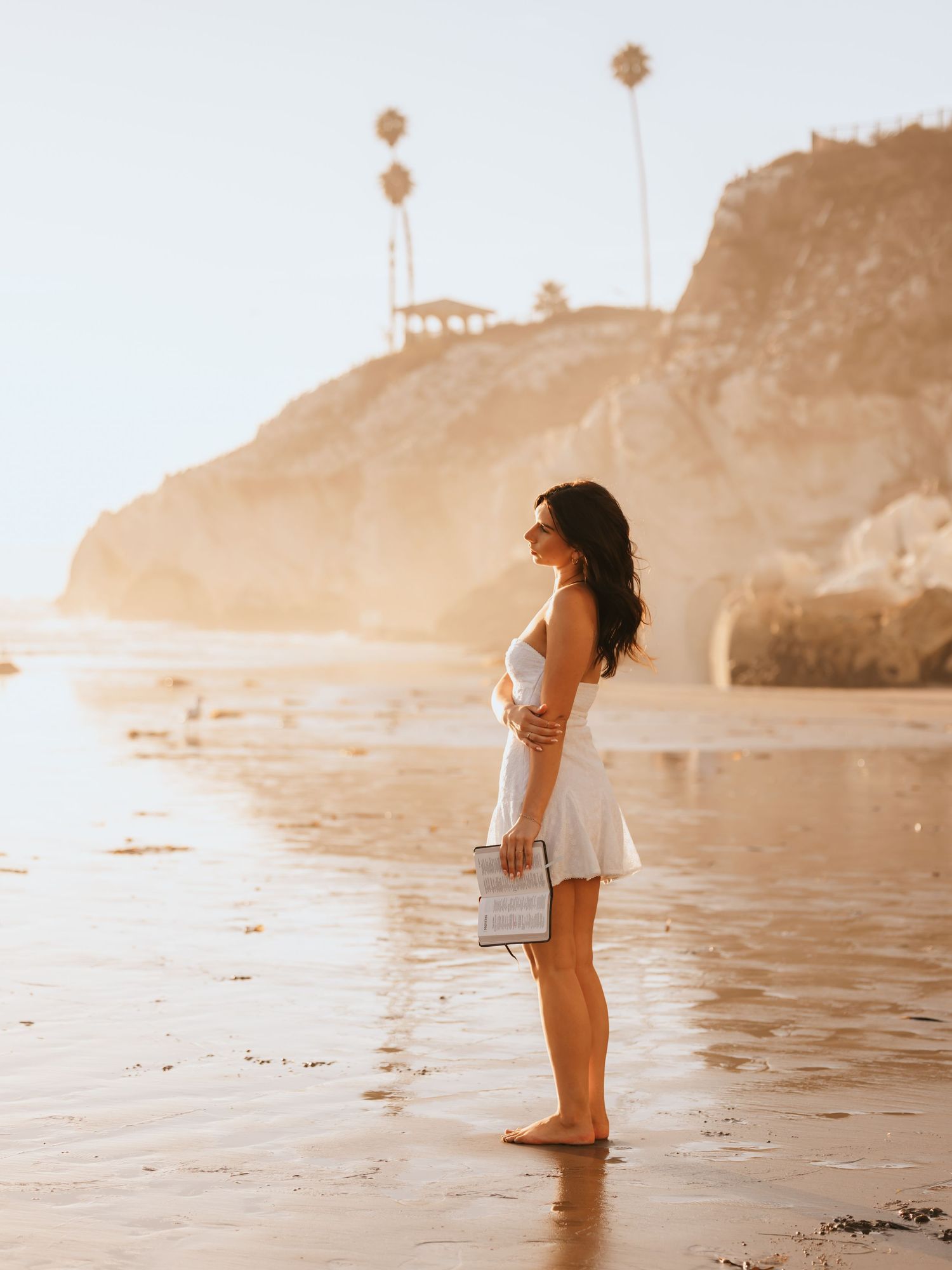 A barefoot woman in a white dress holds a bag on a sunlit beach with rocky cliffs and palm trees.
