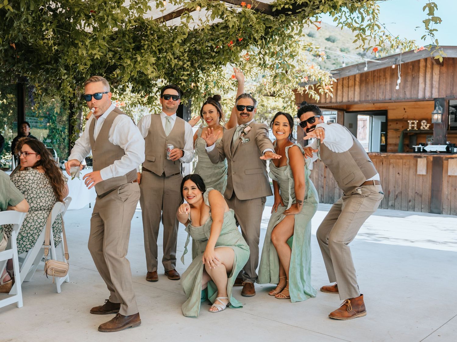 Wedding party posing playfully with drinks under shaded outdoor venue in sage and tan attire.