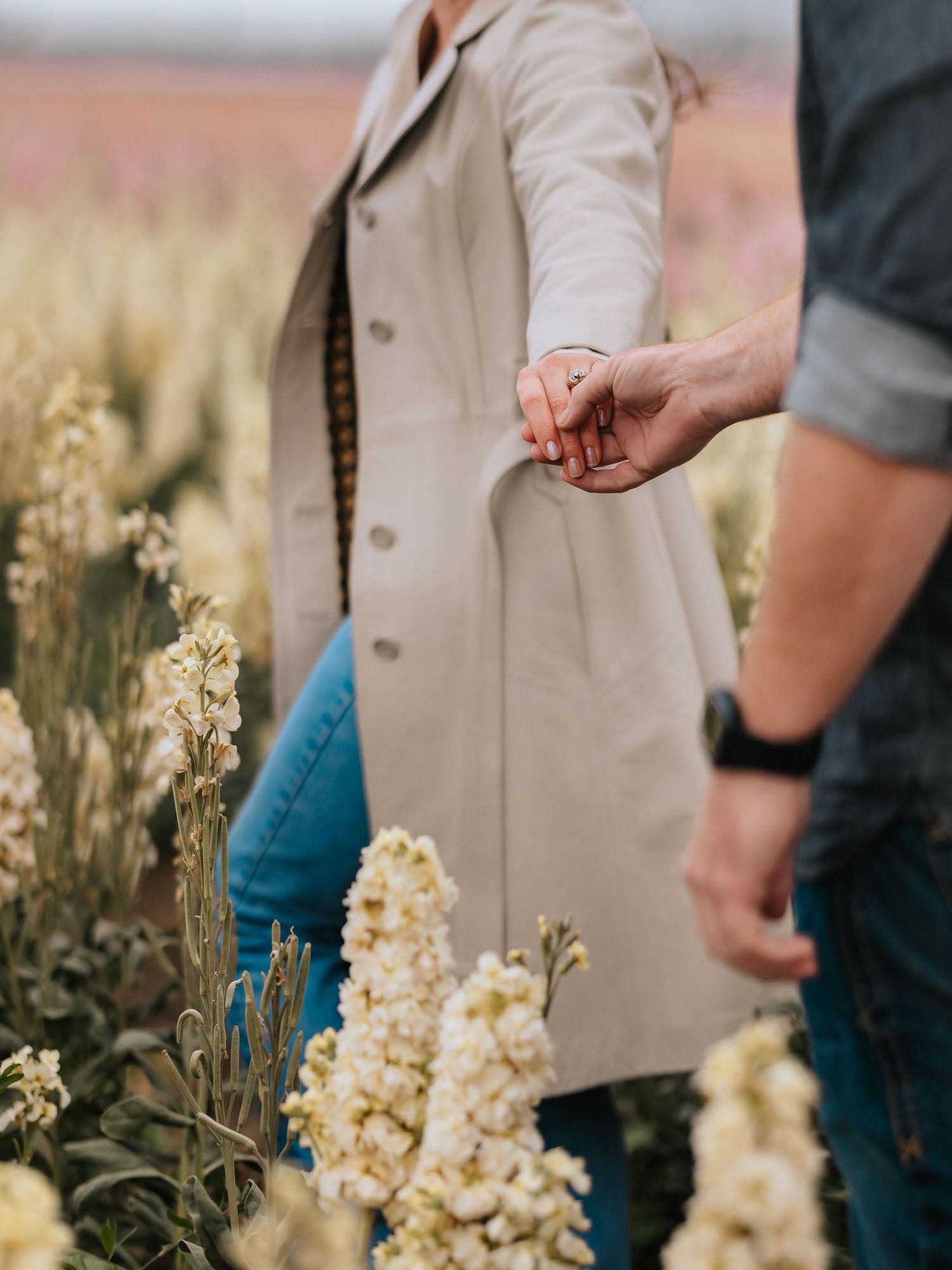 Close-up of two people holding hands in a field of cream-colored flowers with one person wearing a beige coat.