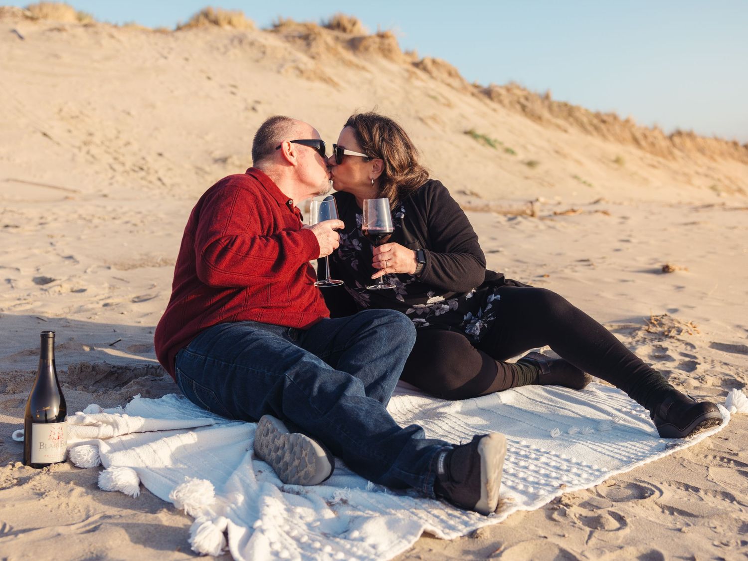 A couple shares a romantic picnic with wine on a sandy beach during sunset.