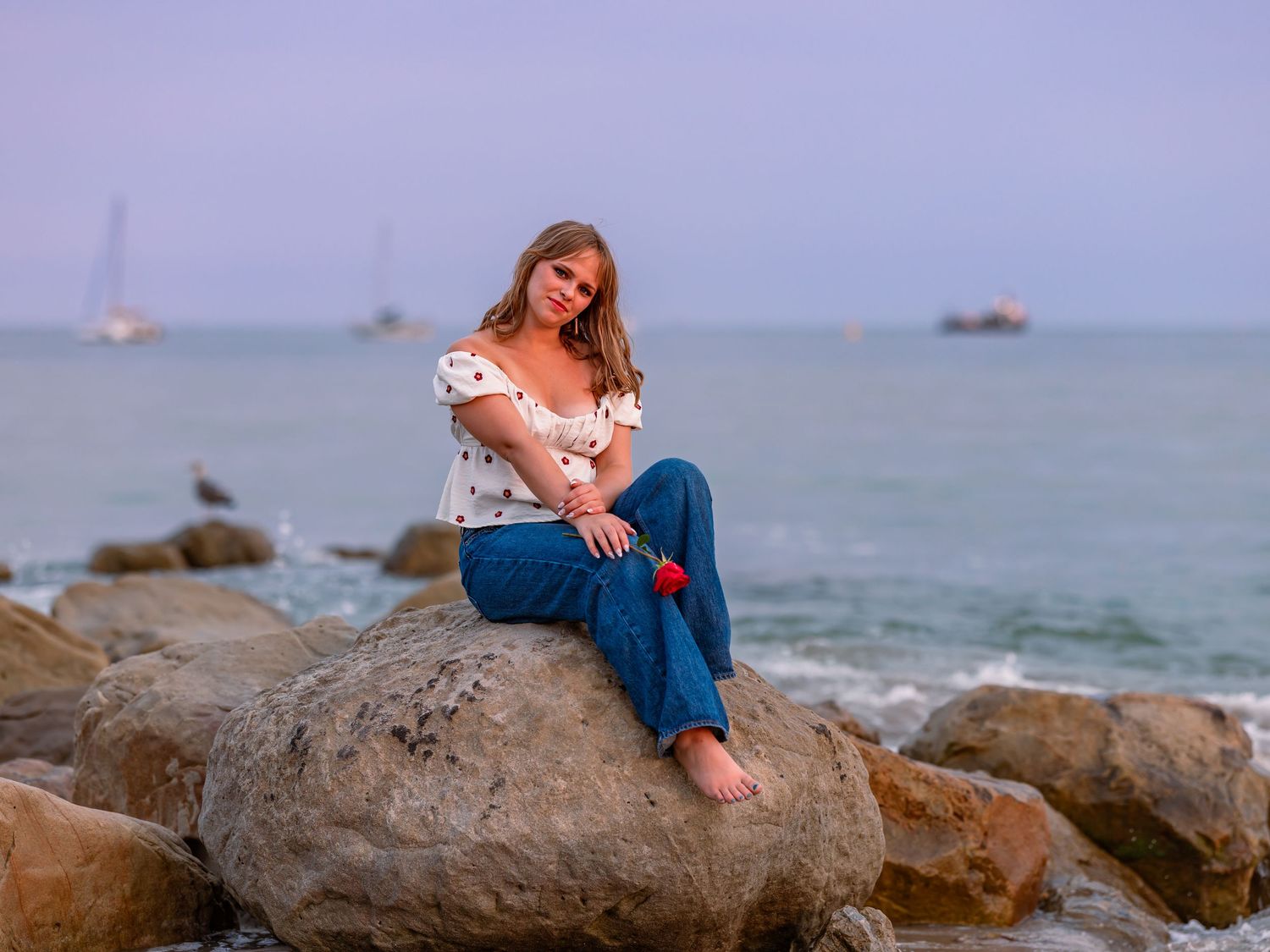 A young woman in jeans and white off-shoulder top sits barefoot on coastal rocks with sailboats visible in the sea behind her.