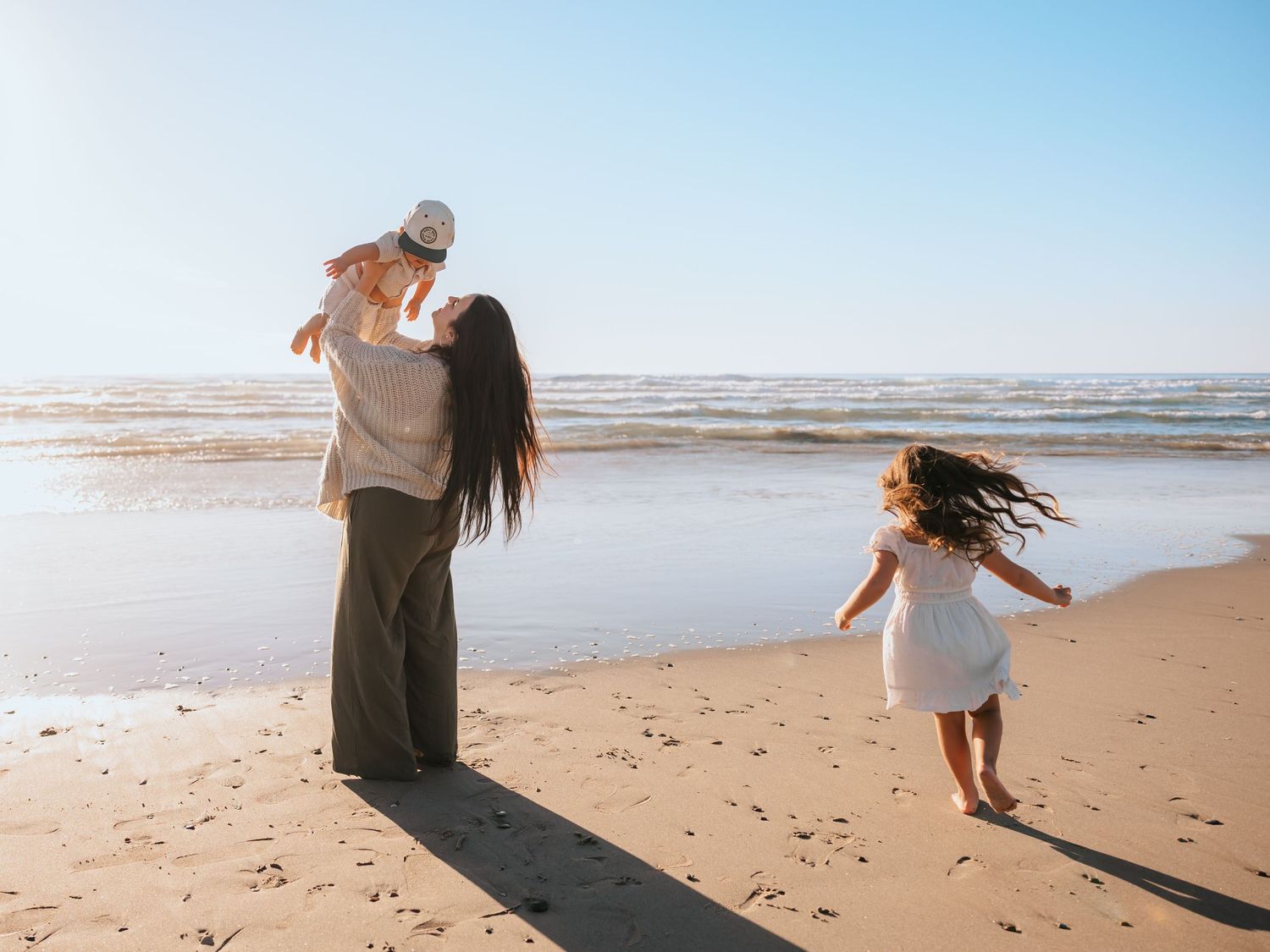 A mother lifts her baby while a young girl runs toward the ocean on a sunny sandy beach.
