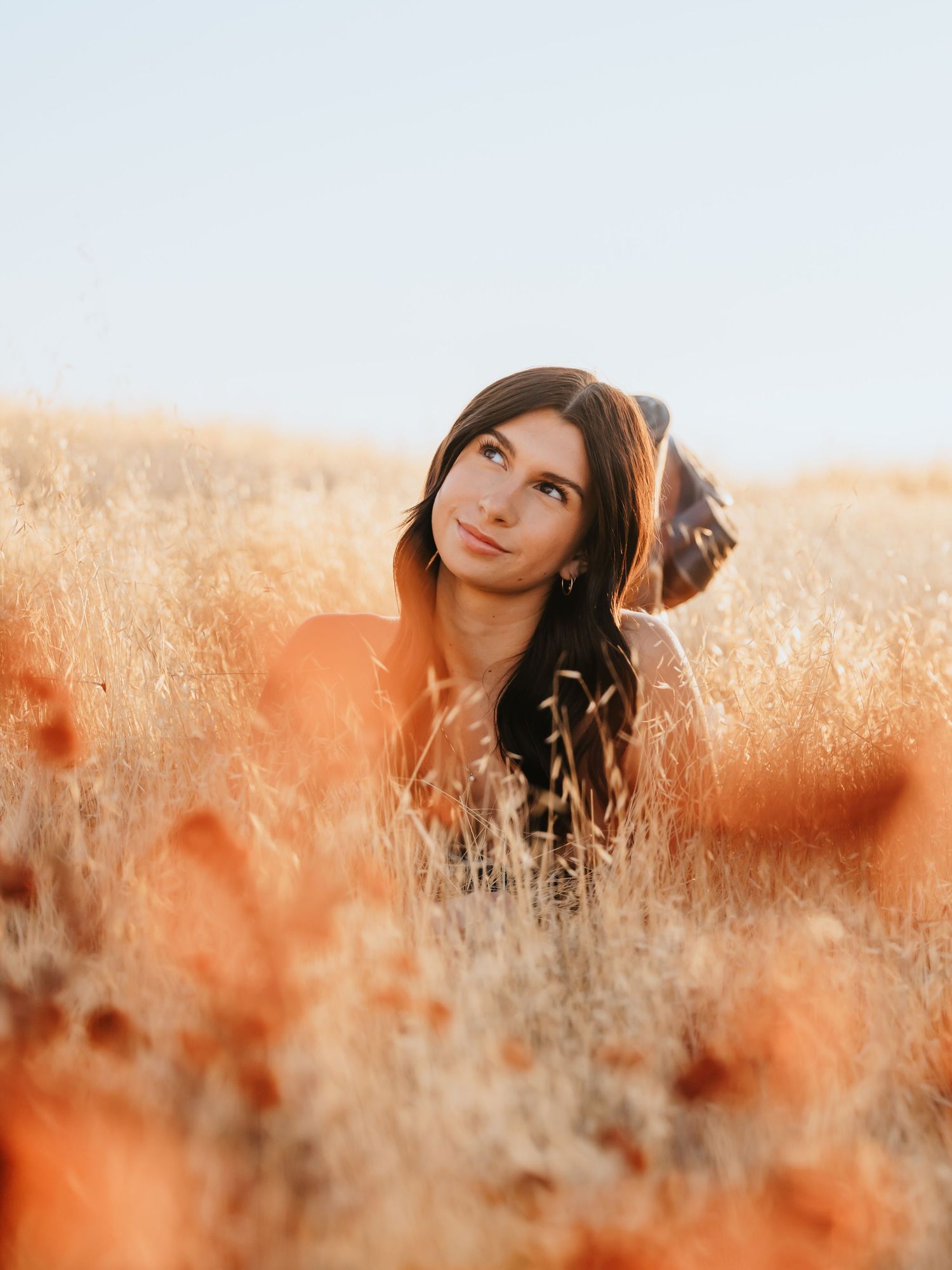 Young woman with dark hair sitting in golden wheat field, gazing upward dreamily in warm autumn sunlight.