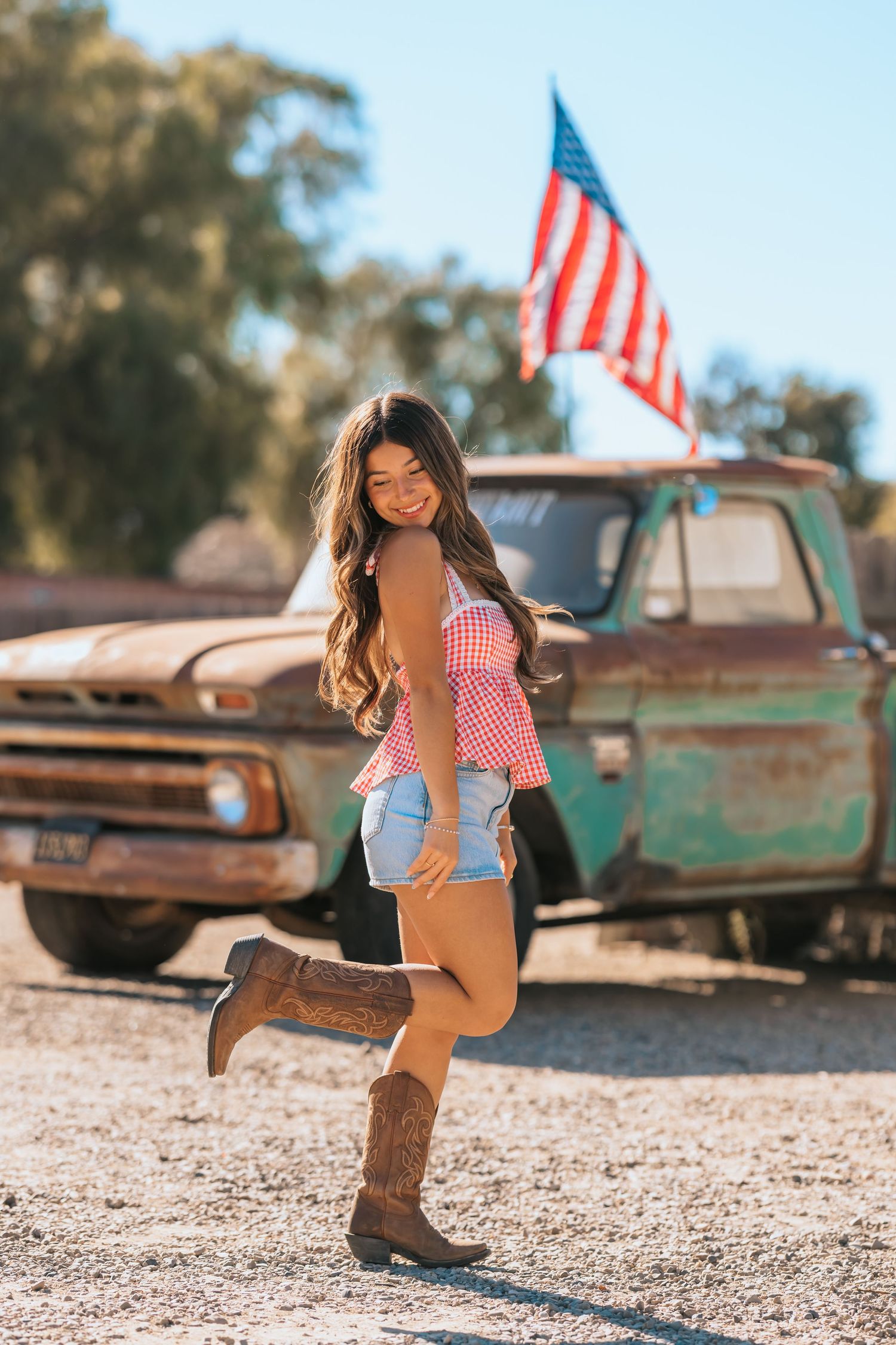 Smiling woman in gingham top and denim shorts posing by vintage truck with American flag.