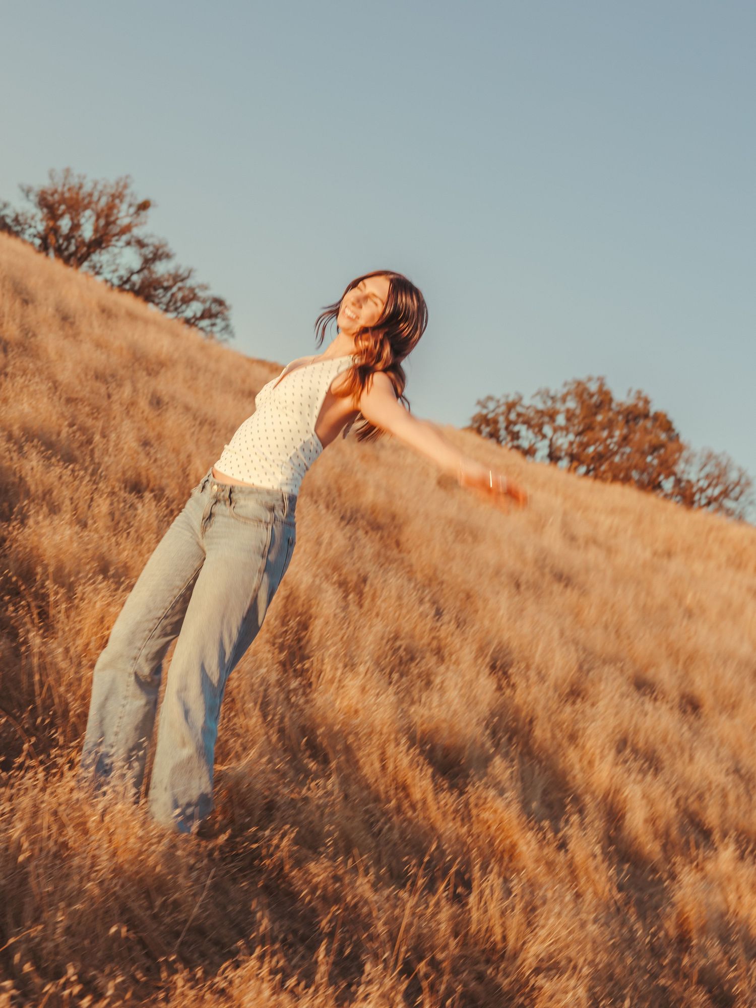 Woman in white top and jeans stands on golden hillside with arms outstretched, basking in warm sunset light.