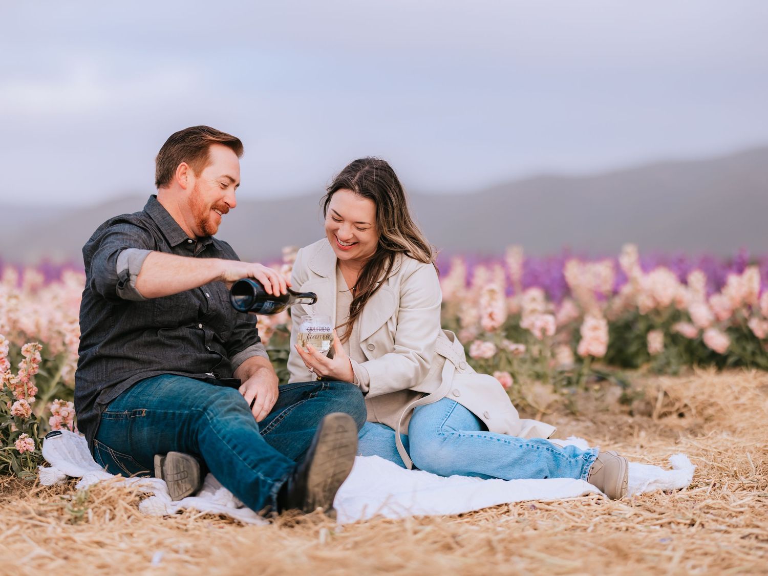 Couple sitting on picnic blanket sharing wine amid wildflowers with mountains in background at sunset.