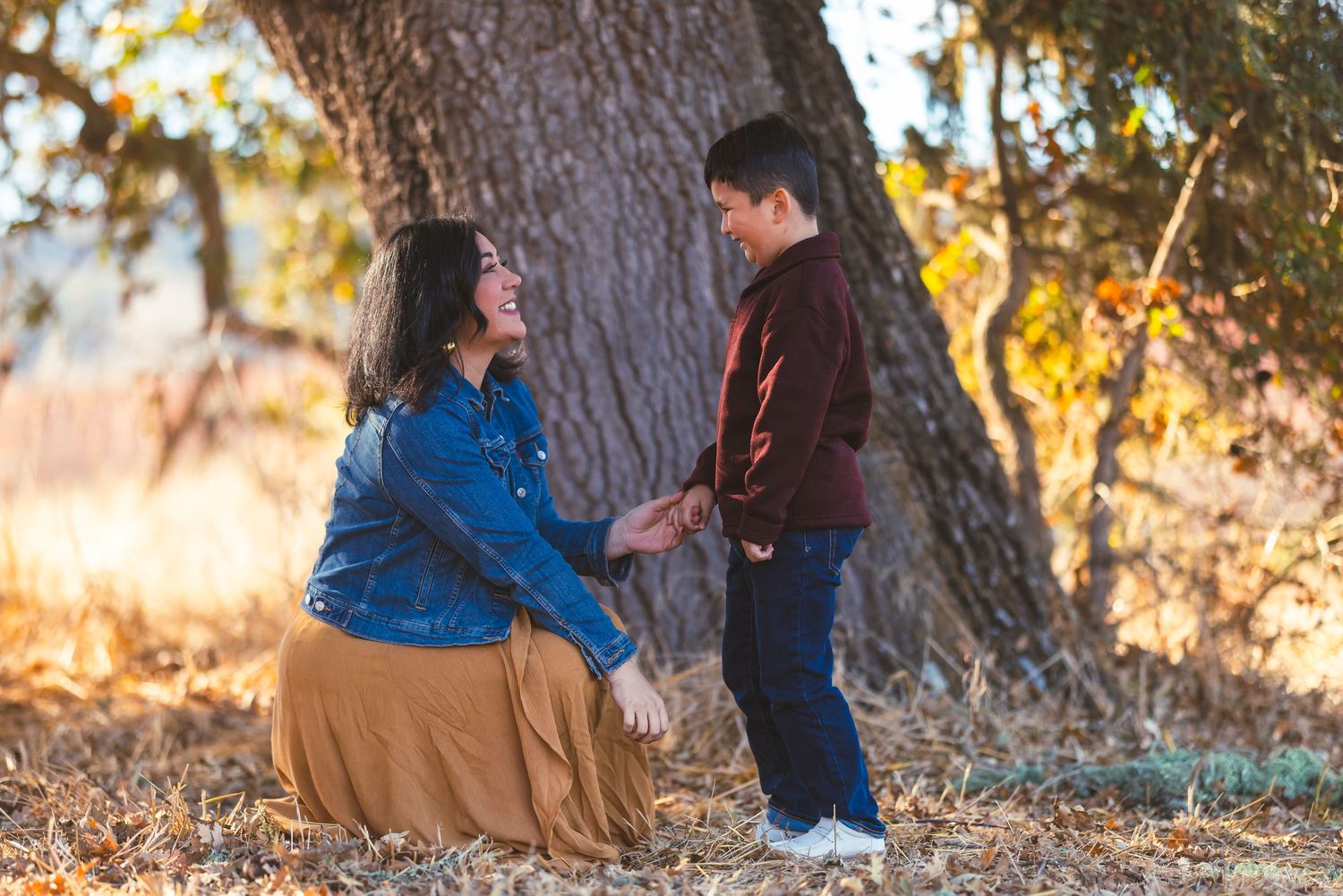 A mother kneels to hold hands with her young son outdoors among autumn leaves near a large tree.