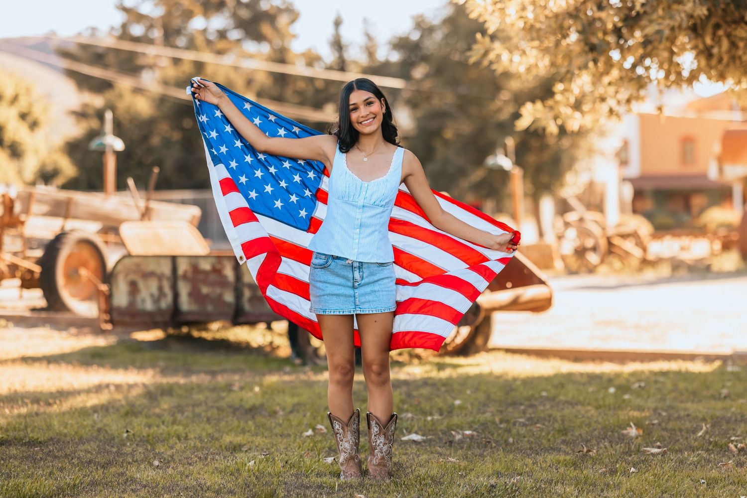 Young woman holding an American flag outdoors, wearing denim shorts and cowboy boots on a sunny day.